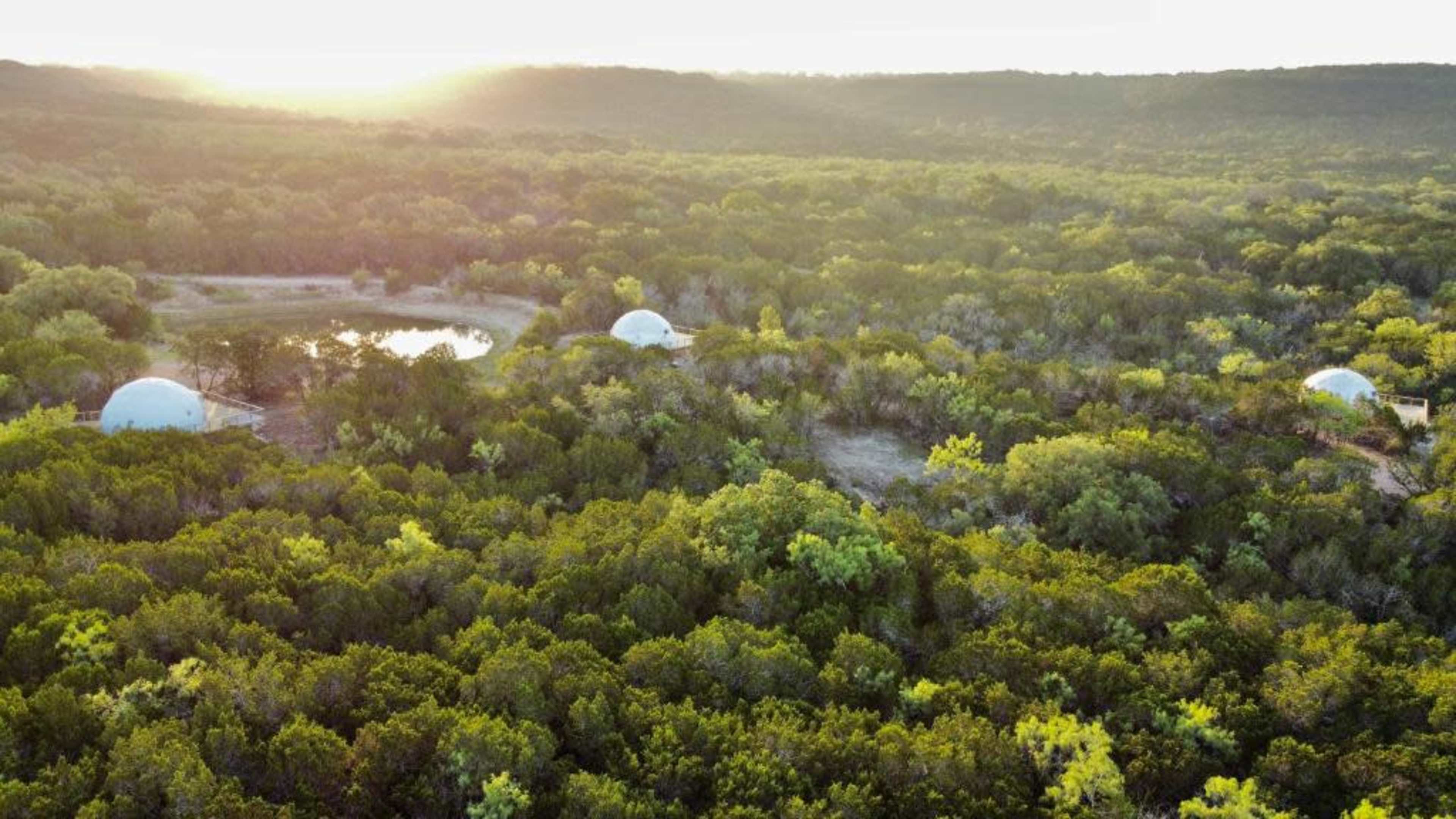 A landscape with domed structures nestled among lush greenery, surrounded by trees and a small body of water in the background.