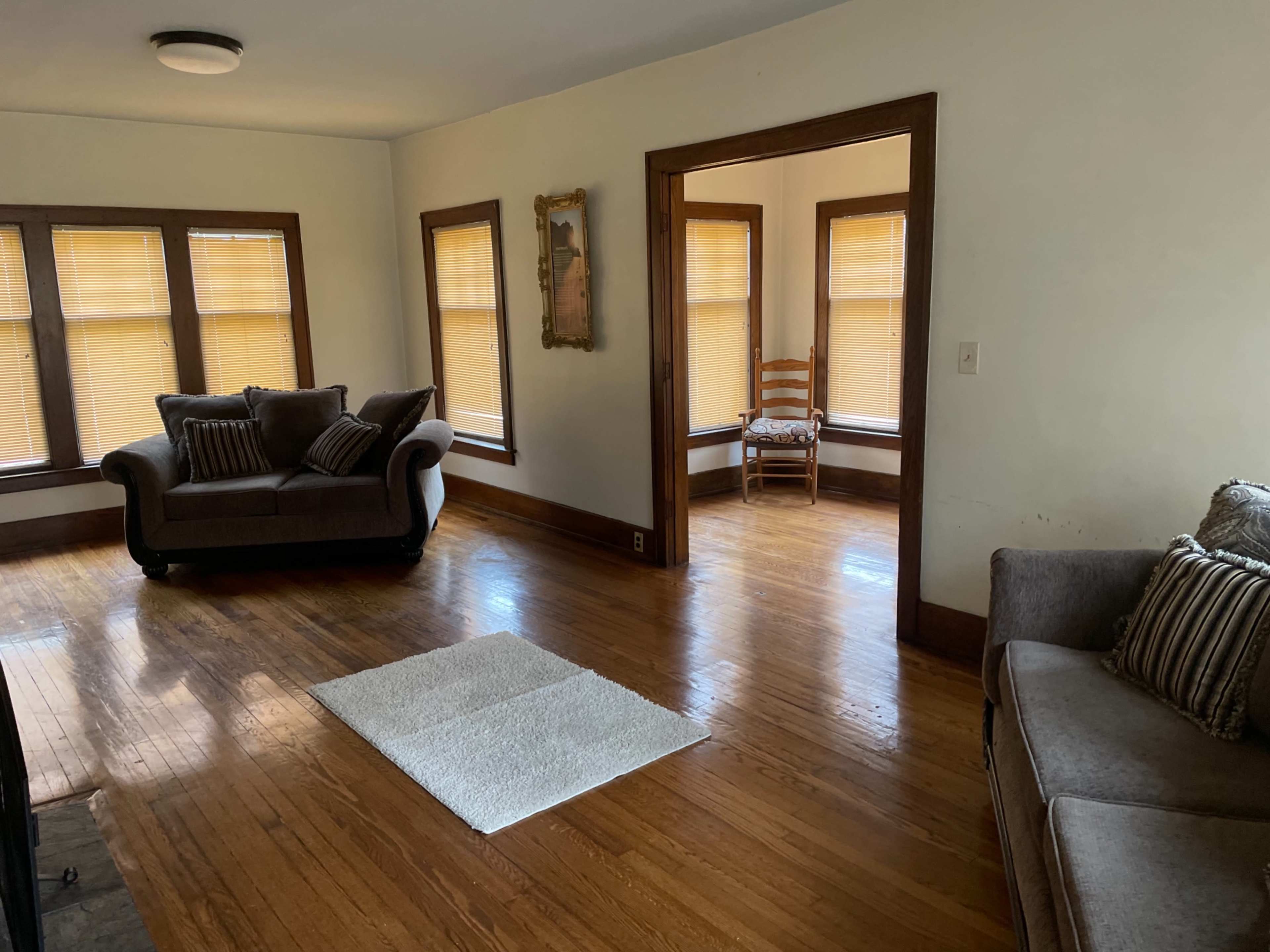 A living room features two sofas, a coffee table on a rug, and large windows with shades, leading to an adjacent room with a chair.