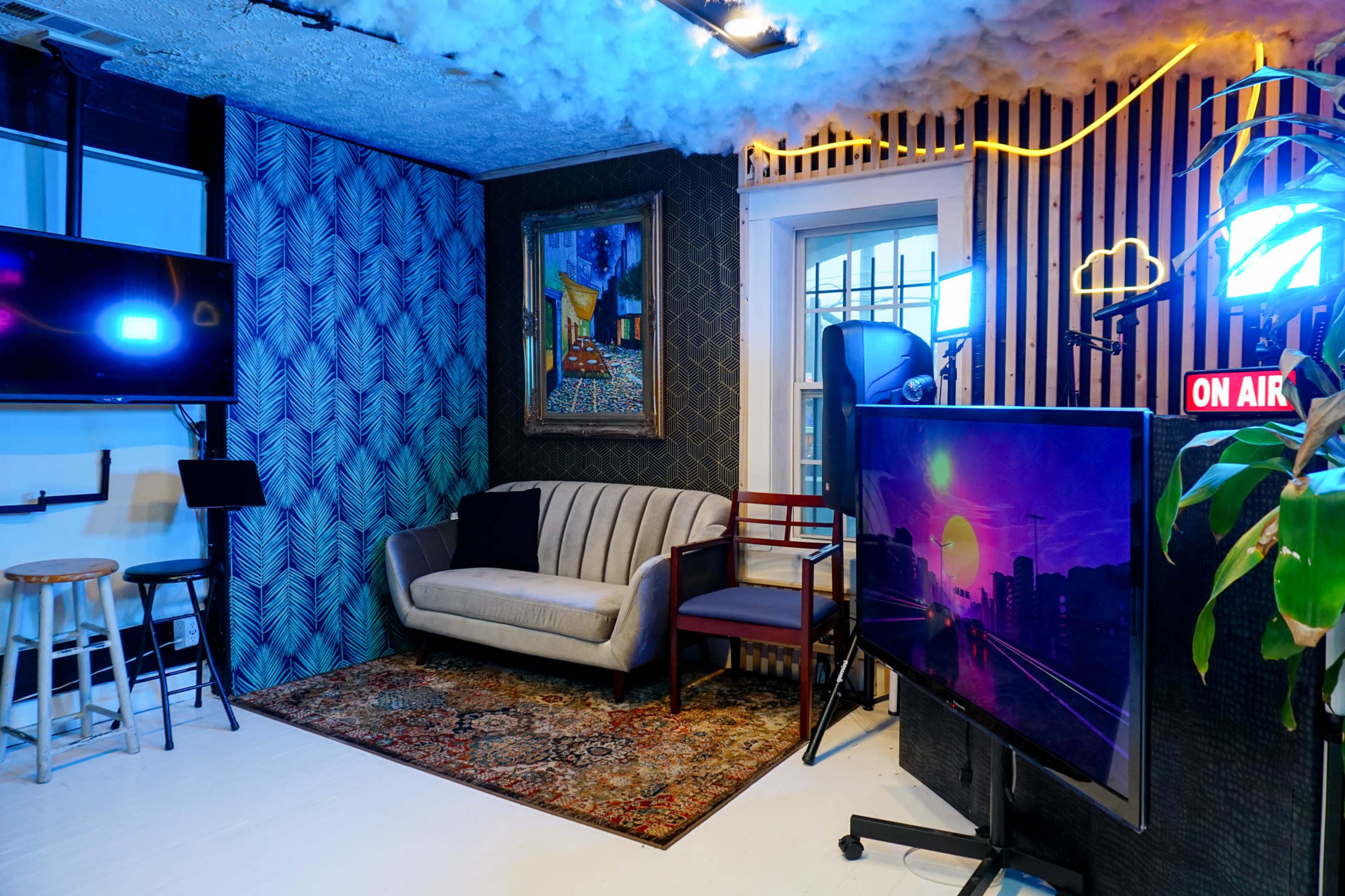 The image shows a cozy studio space featuring a grey sofa, decorative walls, and multiple screens with lighting setups.