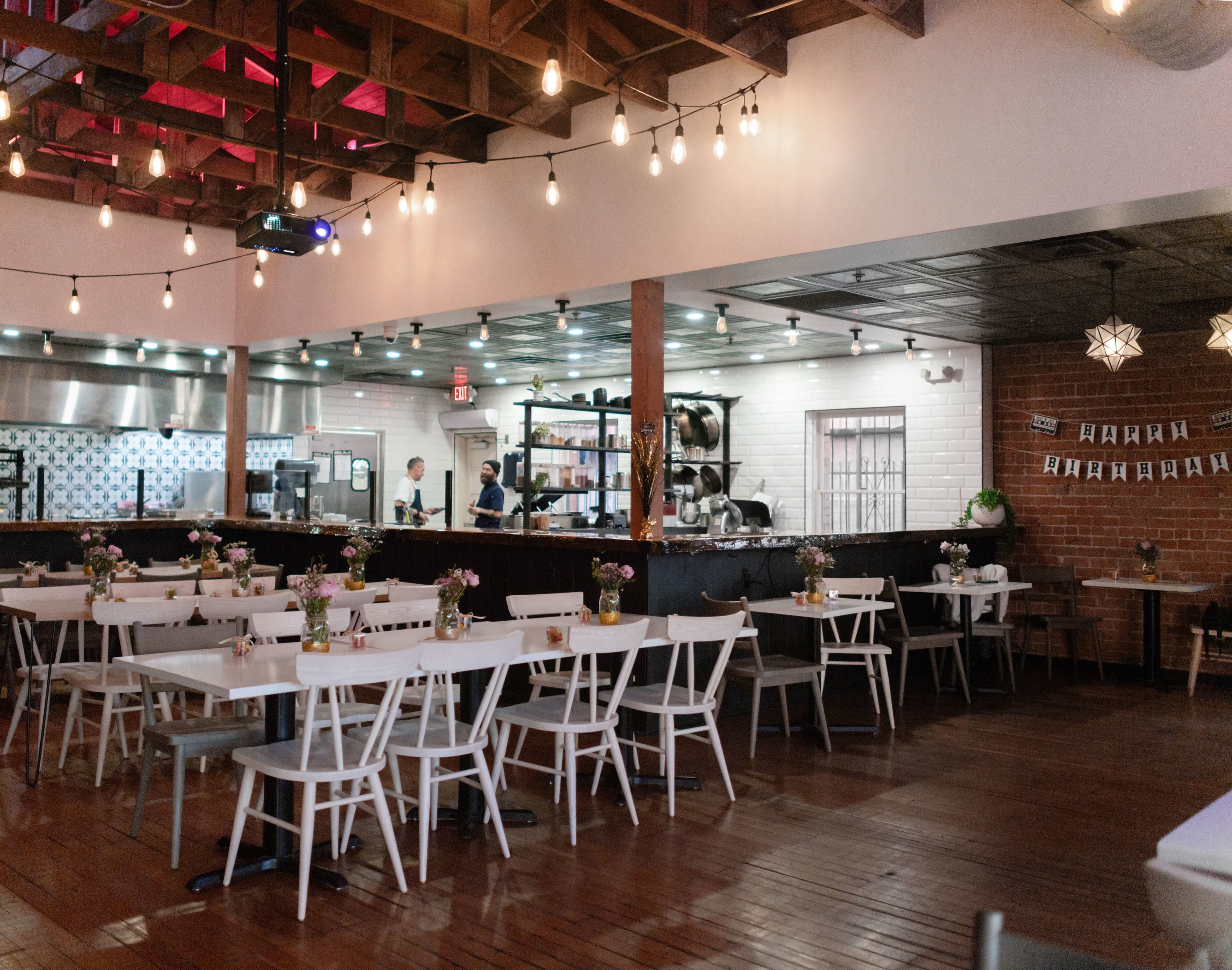 The image shows a restaurant interior with wooden floors, a row of tables with flowers, and an open kitchen area in the background.