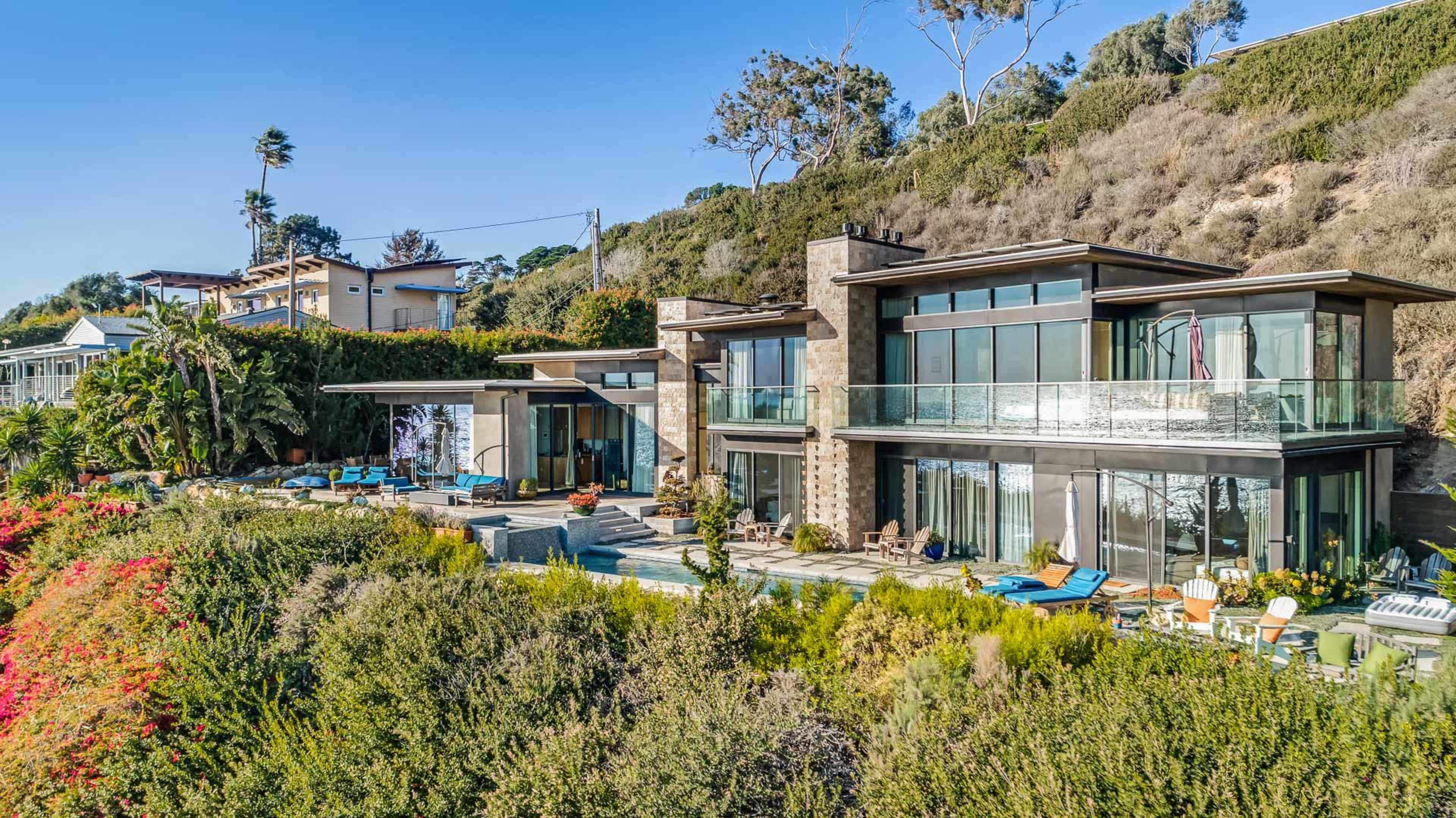 The image shows a modern hillside house with large glass windows surrounded by greenery and outdoor seating areas.