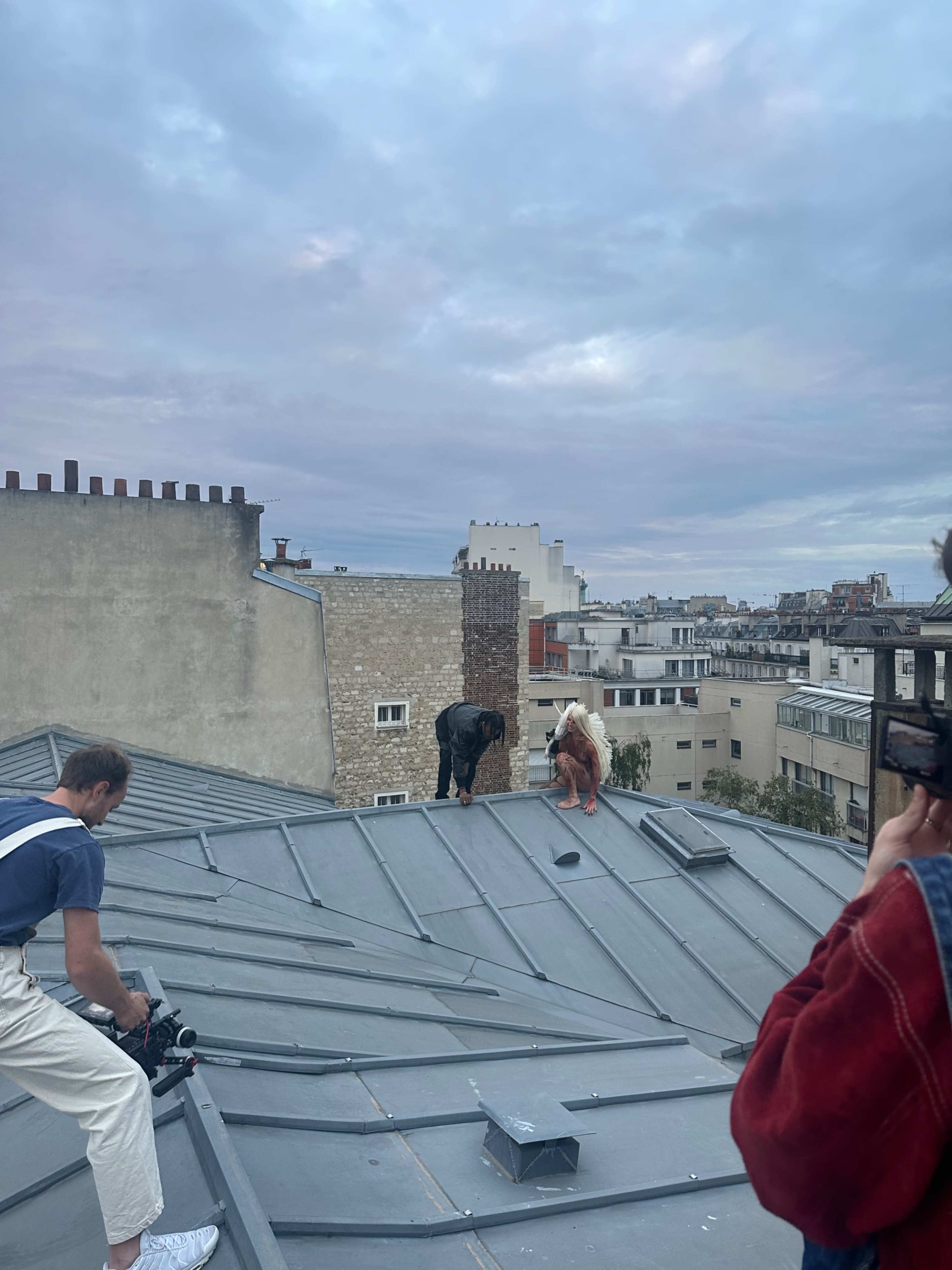 Two individuals are climbing on a metal roof amidst an urban environment, while a third person records the scene with a camera.
