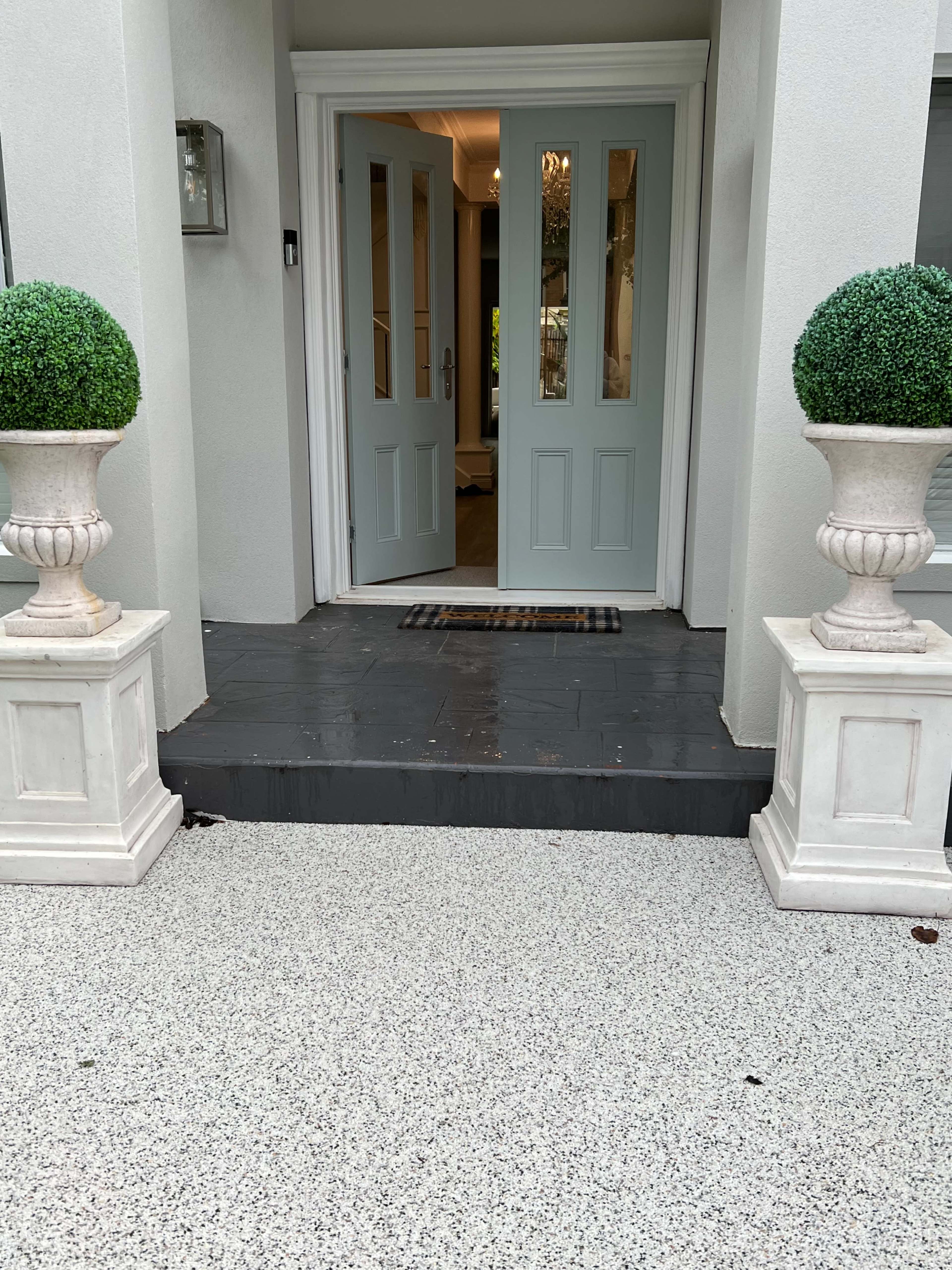A front entrance with light blue double doors, flanked by white planters containing spherical topiaries, on a speckled gray concrete surface.