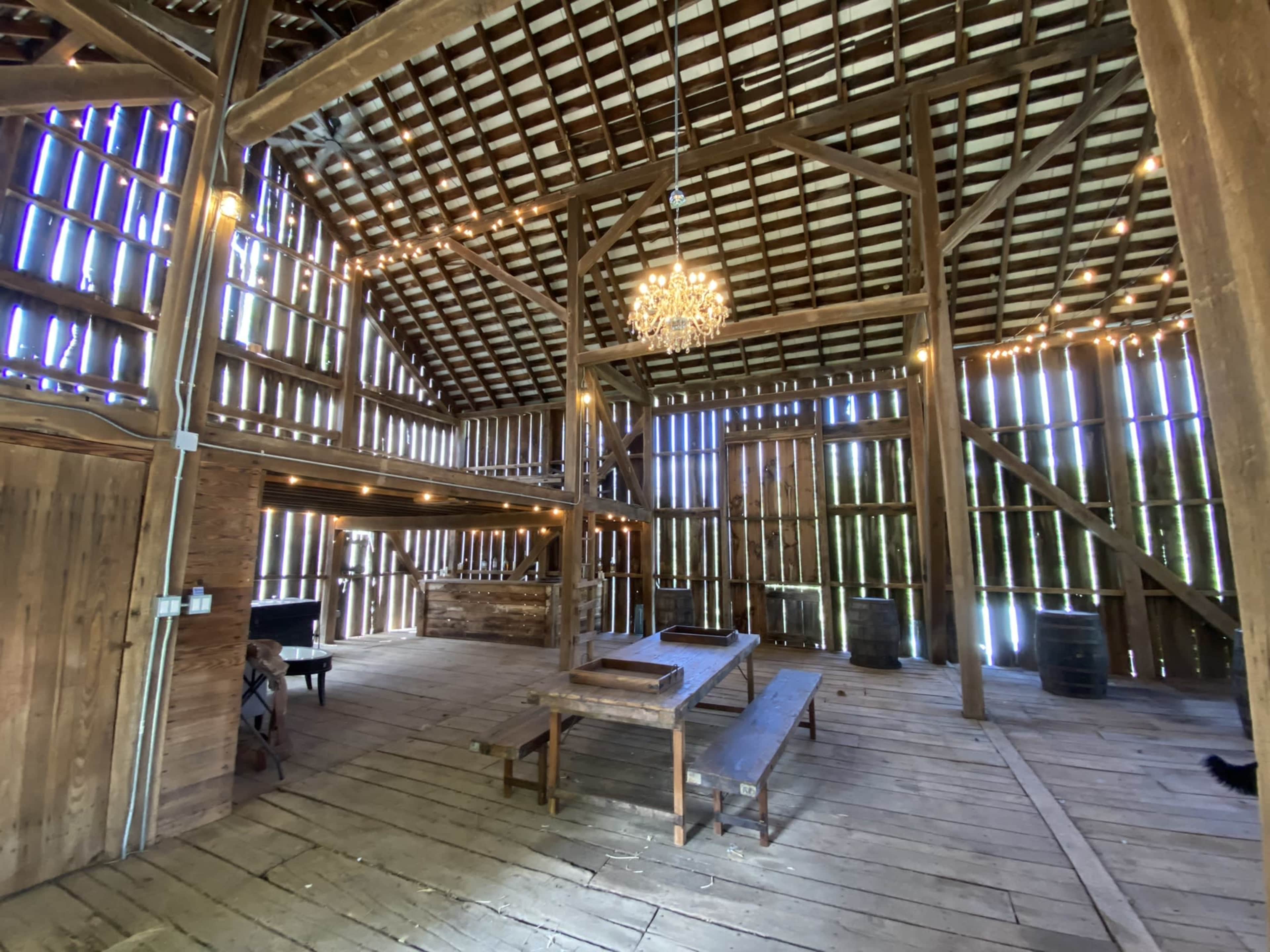 The interior of a spacious barn with wooden beams, a high ceiling, a chandelier, and wooden tables surrounded by rustic walls.