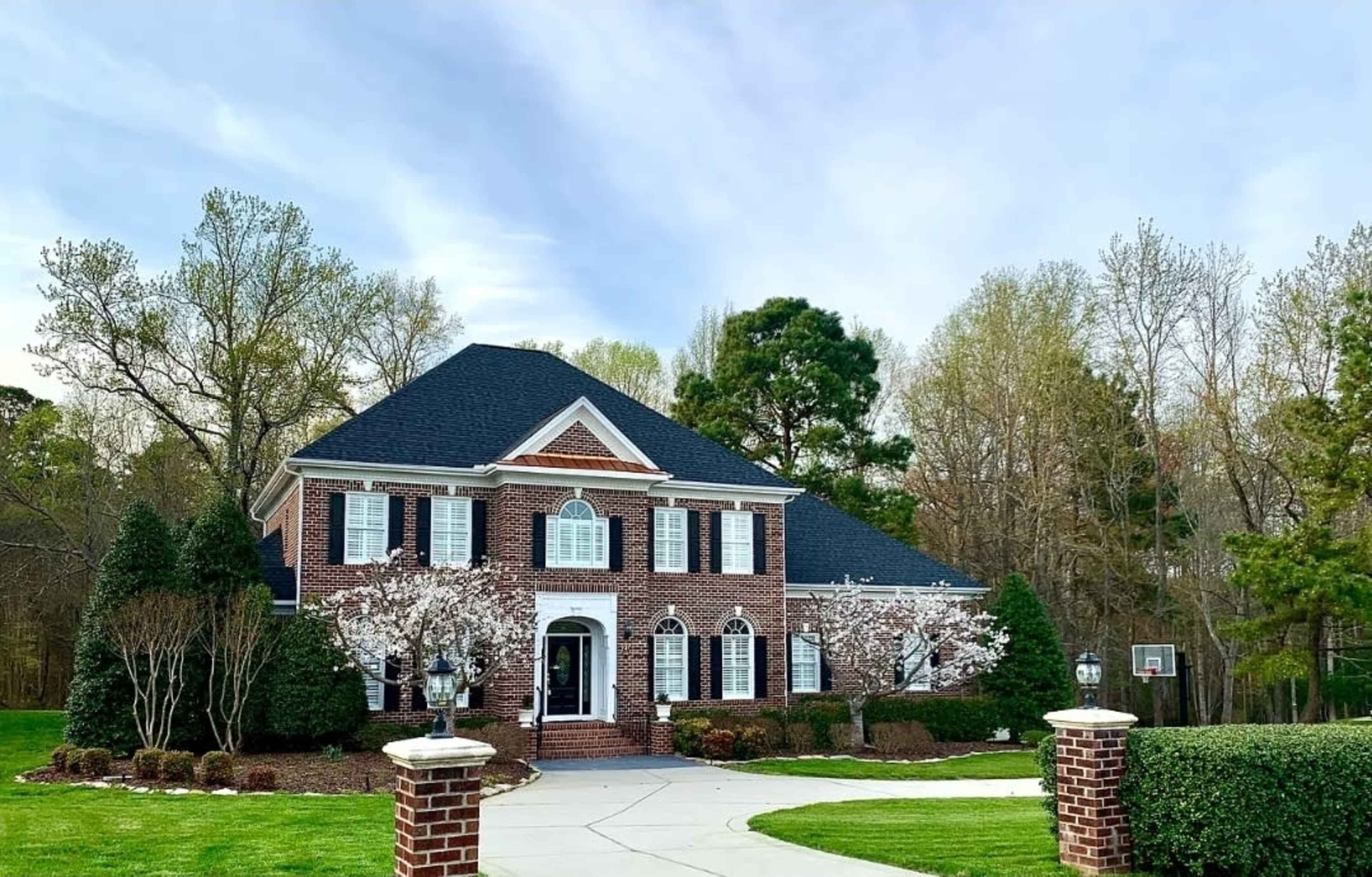 A large brick house with a dark roof and white shutters is set in a landscaped yard with trees and blooming shrubs.