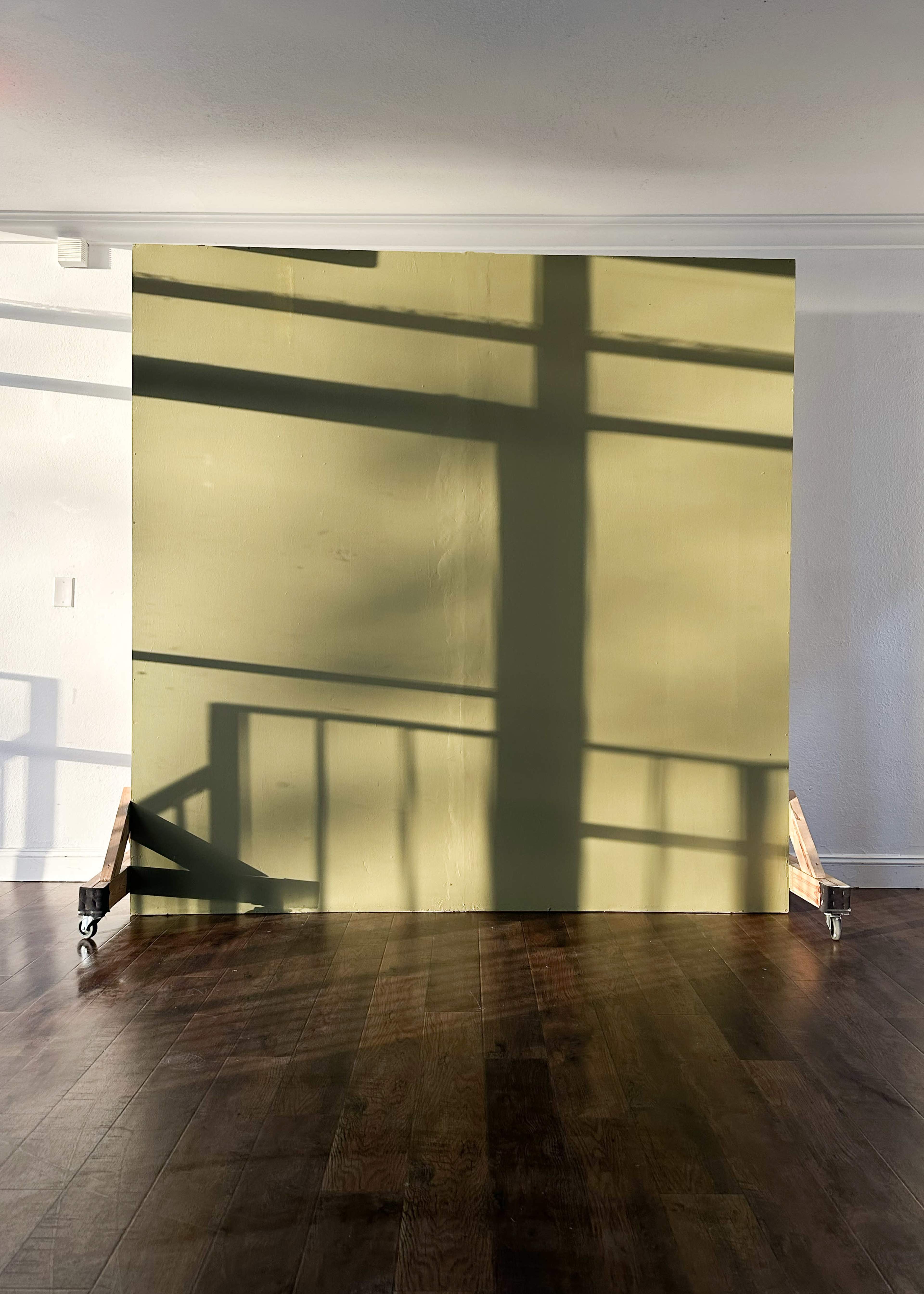 A large green wall stands in an empty room with wooden flooring, casting shadows from a nearby object.