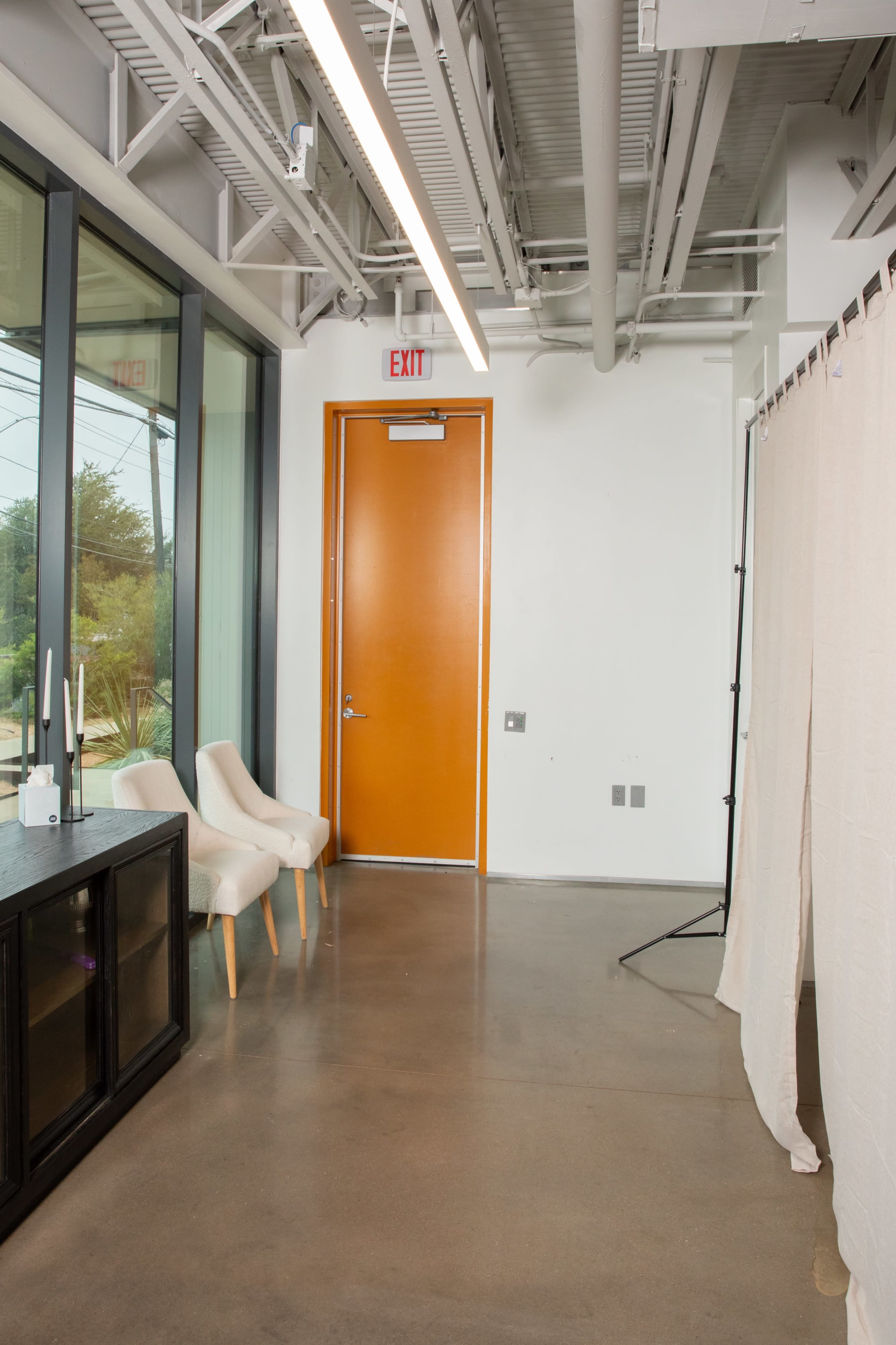 The image shows a modern interior space with two light-colored chairs, a dark cabinet, an orange door, and a privacy curtain on one side.