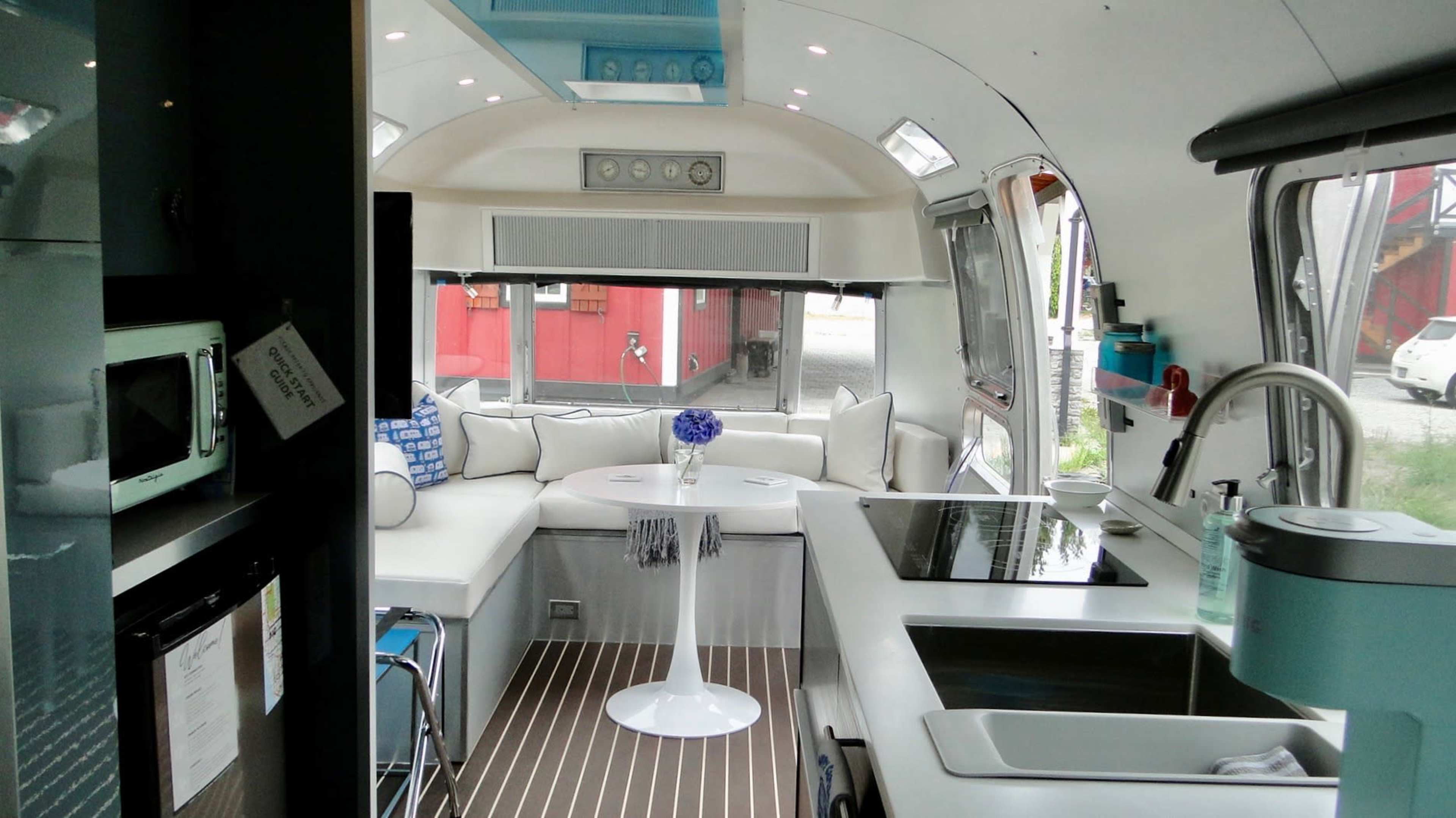 The image shows the interior of a modern, renovated Airstream trailer with sleek furnishings and a kitchen area featuring a stove and sink.