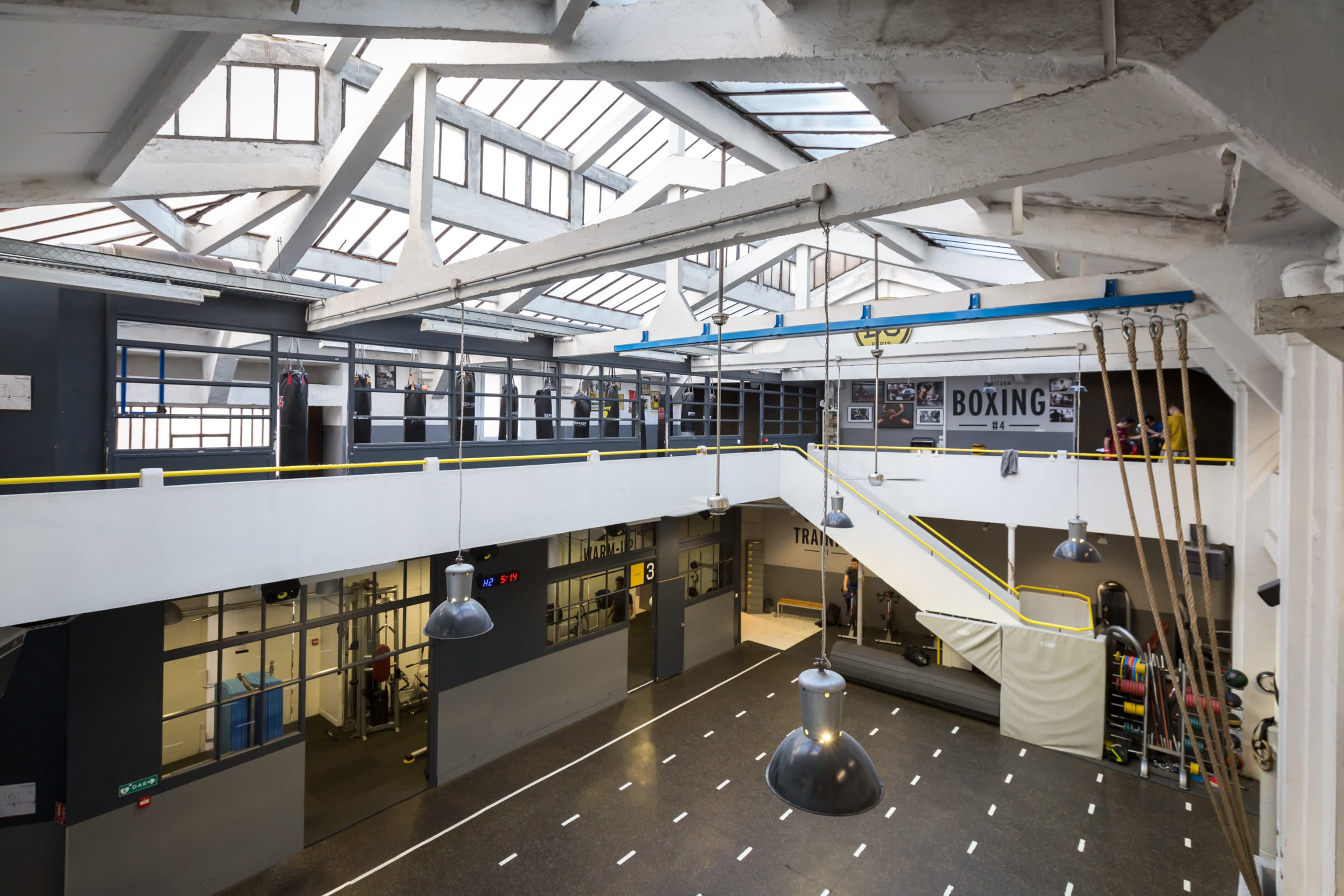 A spacious gym interior with multiple levels, featuring a boxing area, training spaces, and large windows allowing natural light to enter.