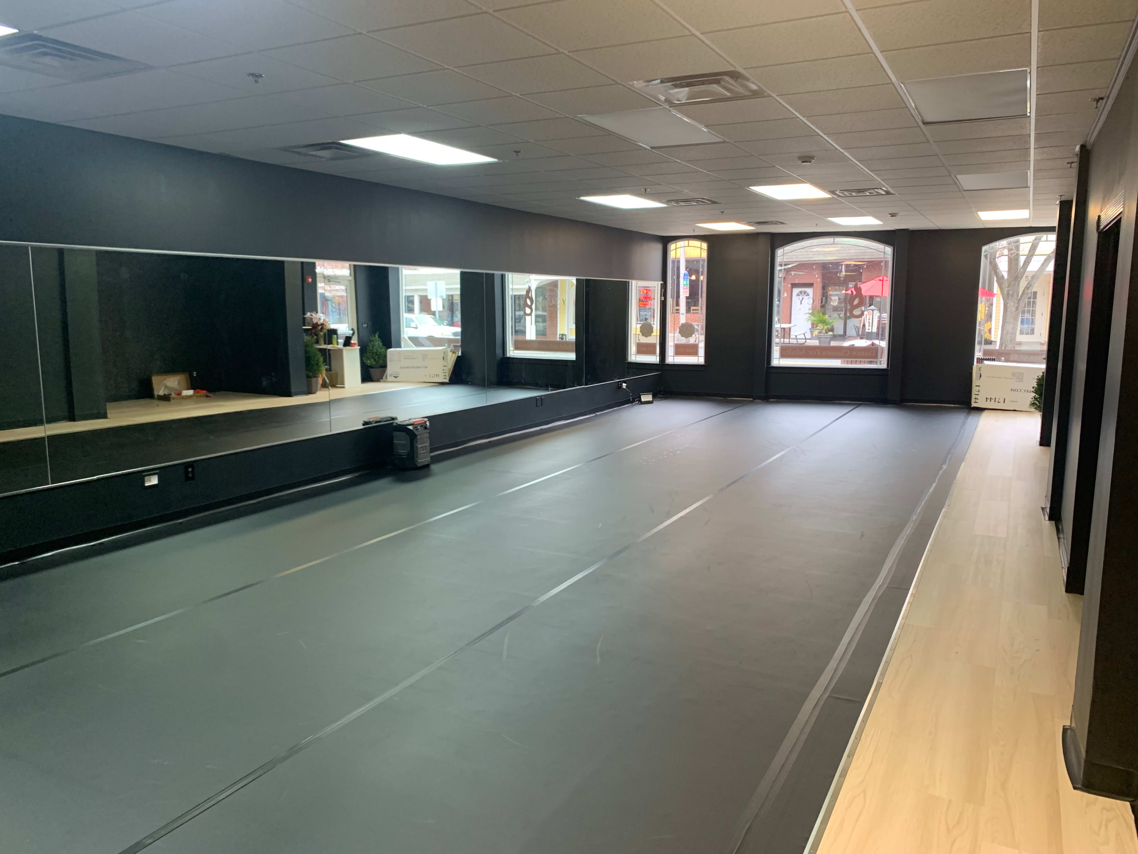 A spacious, empty room with mirrored walls and a black mat flooring, illuminated by natural light from large windows.