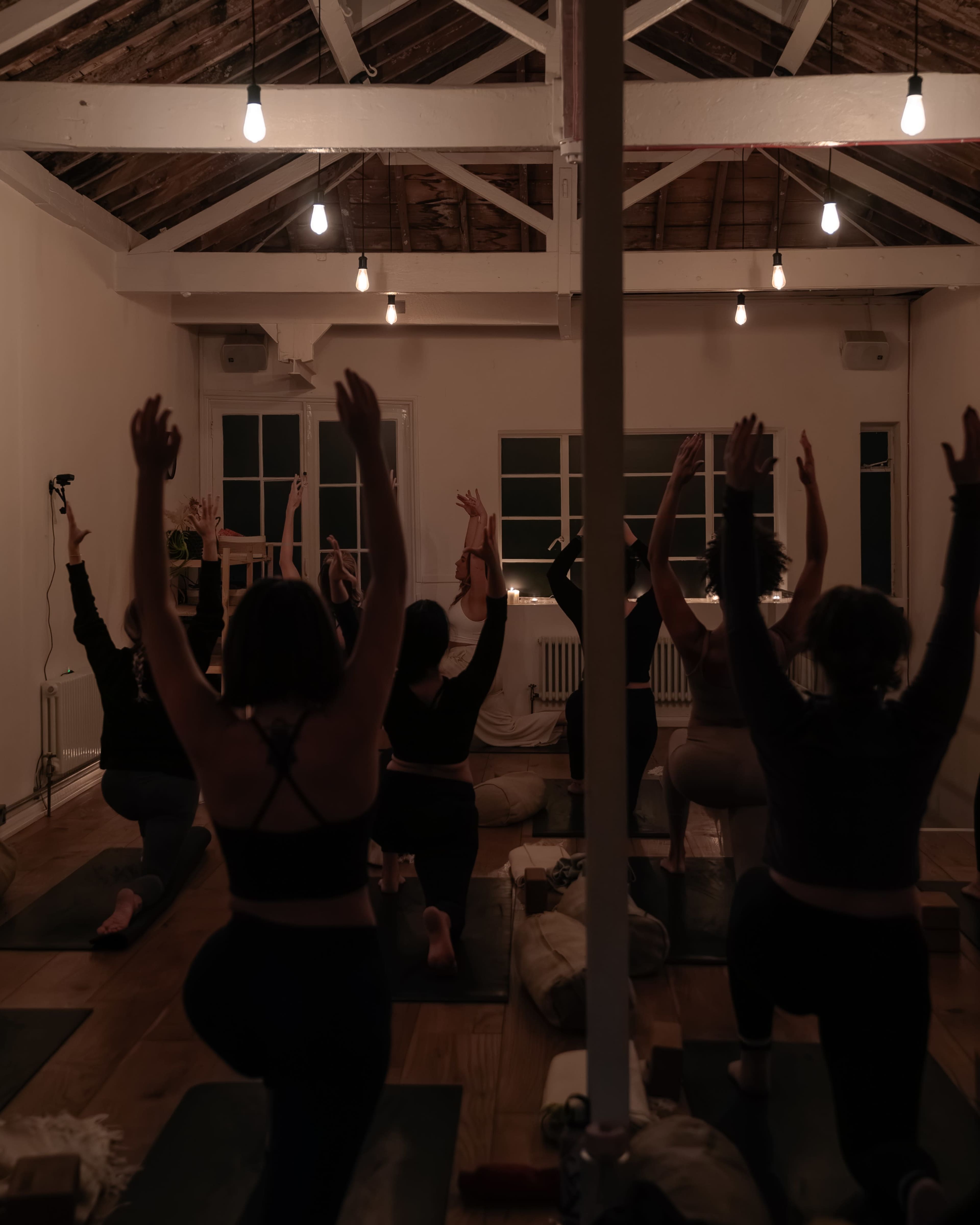 A group of people is practicing yoga in a dimly lit studio with wood beams and low-hanging lights.