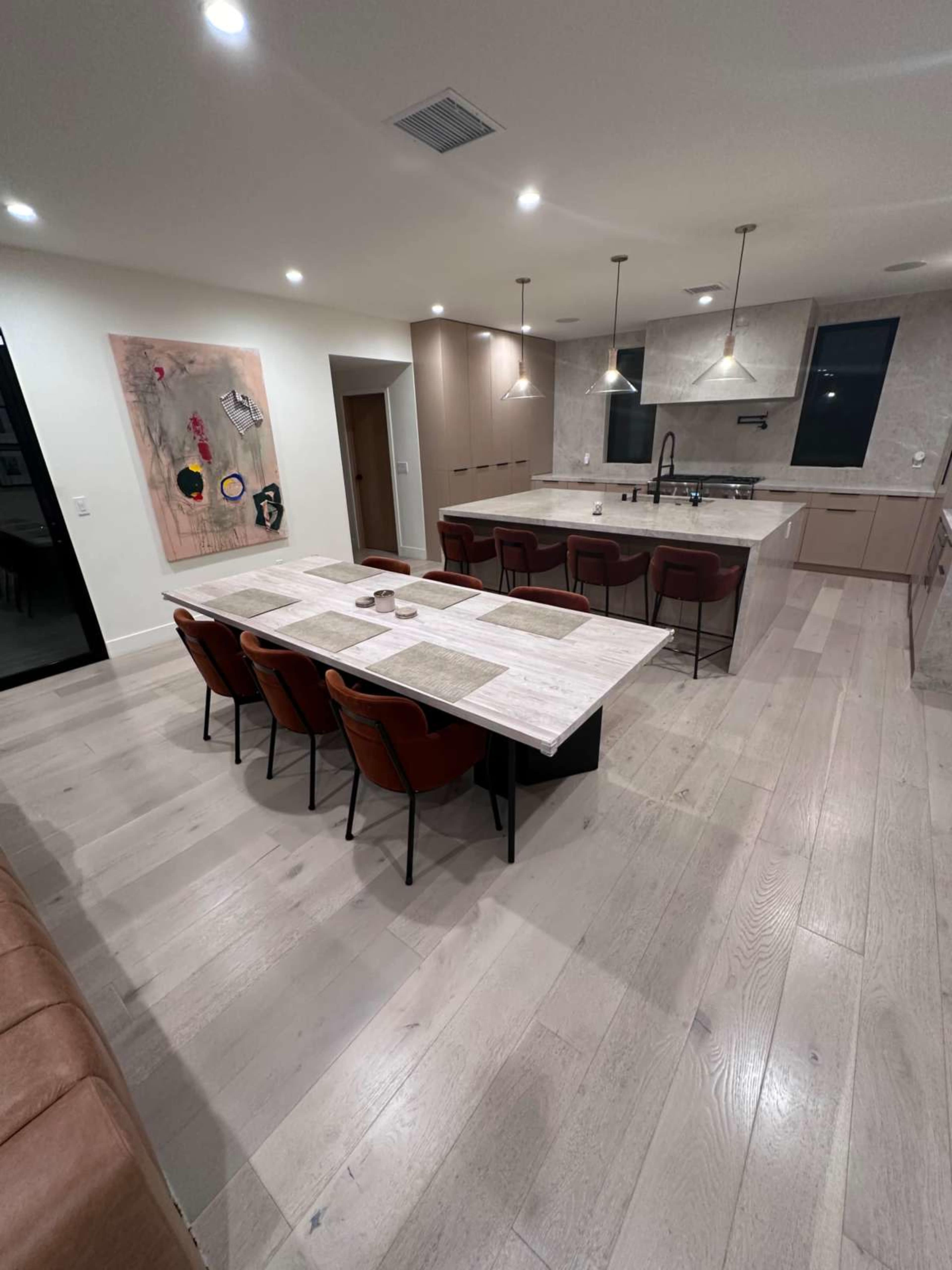 The image depicts a modern kitchen and dining area featuring a large table surrounded by chairs, sleek cabinetry, and pendant lighting above the kitchen island.