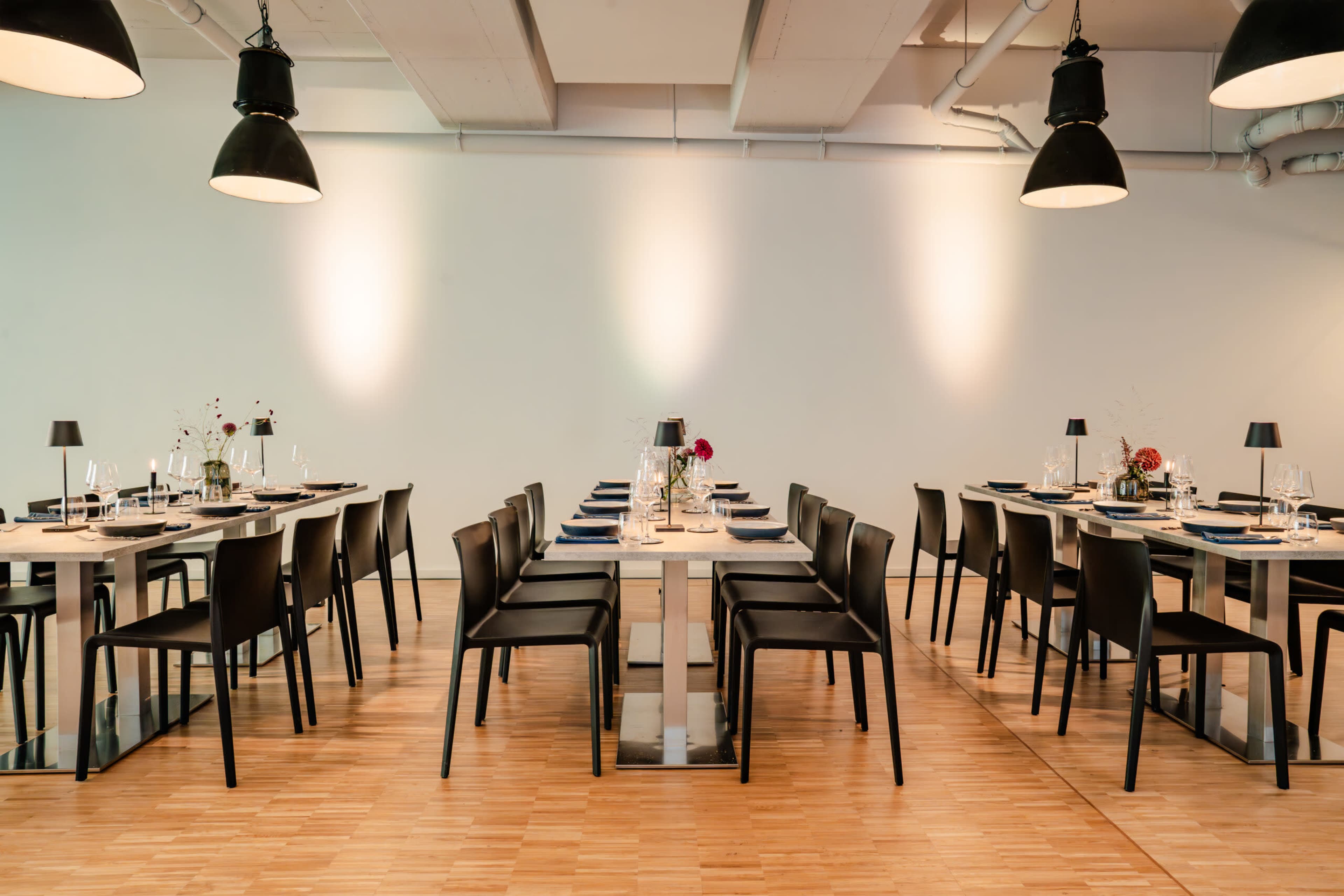 A modern dining room features multiple black chairs arranged around wooden tables, illuminated by pendant lights hanging from the ceiling.