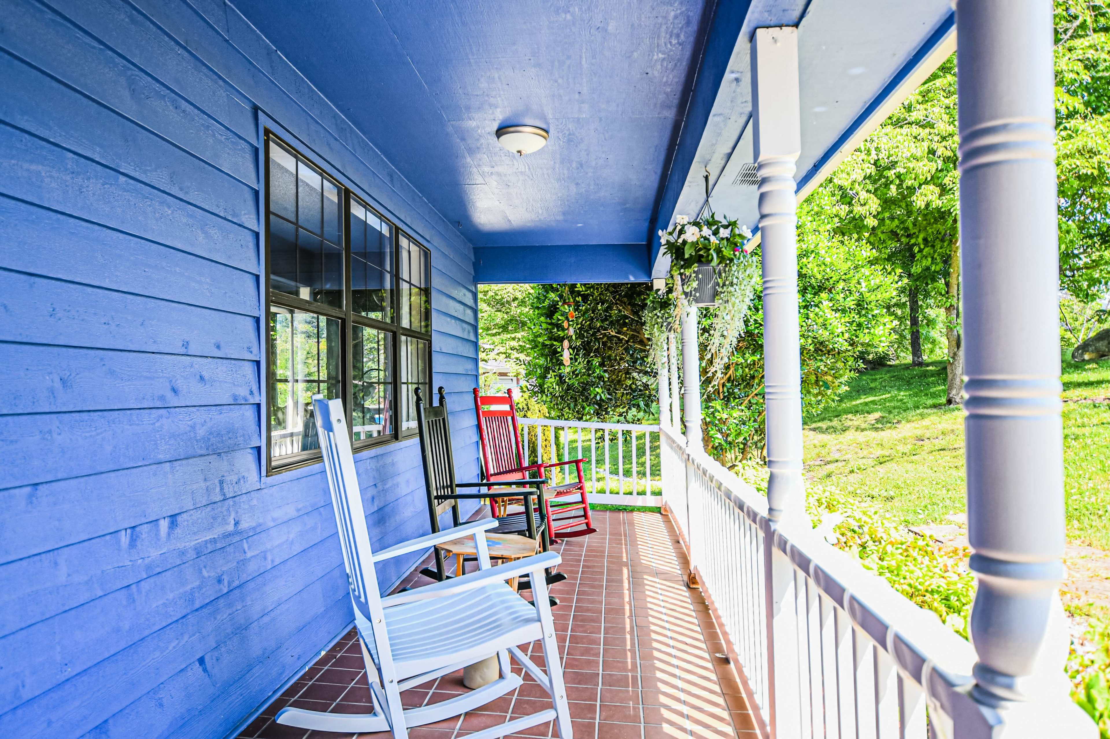 A blue house porch features two rocking chairs, one white and one red, with a view of a grassy area.
