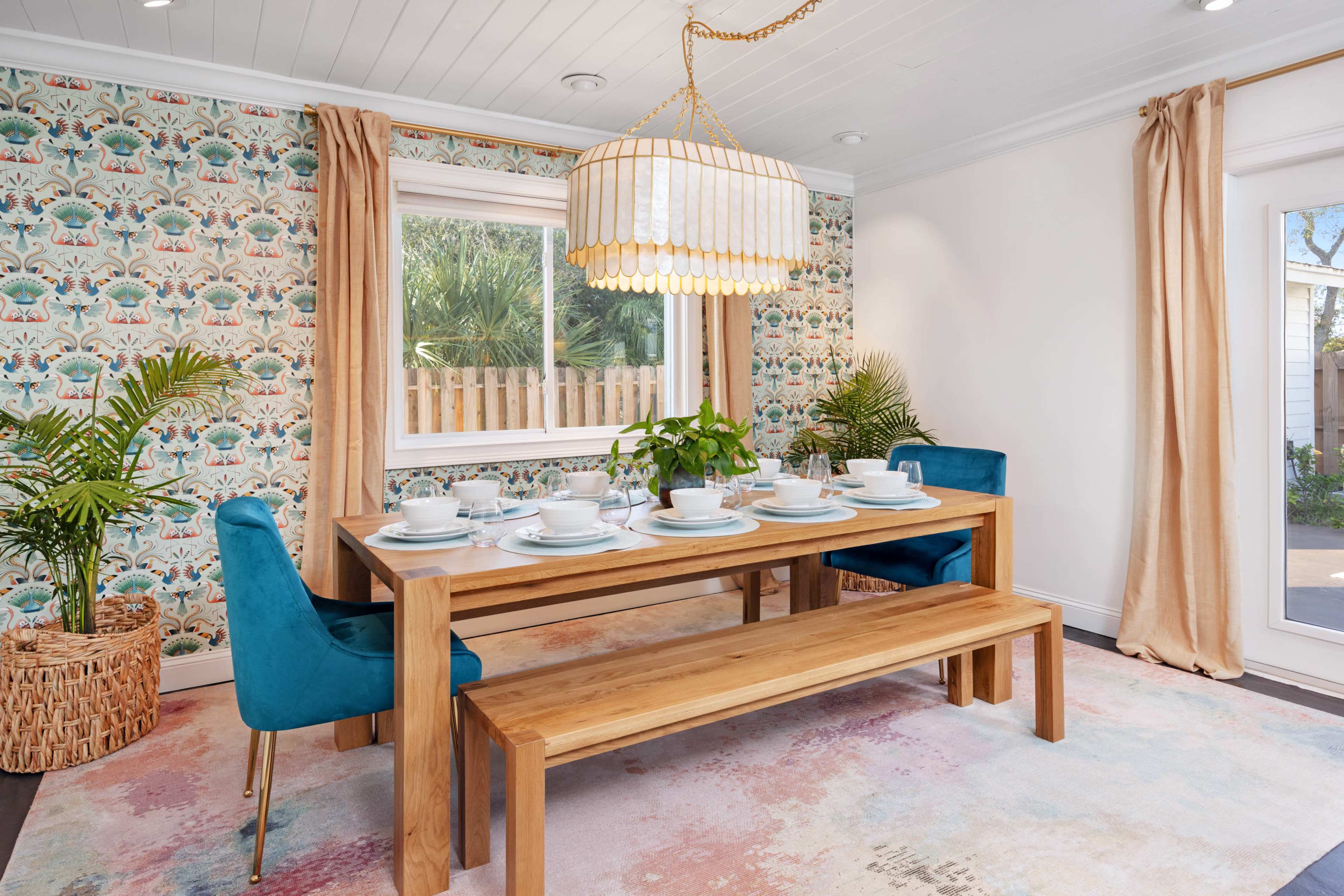 A dining room features a wooden table set for six, surrounded by blue chairs, a long chandelier above, and decorative wallpaper with vibrant patterns.