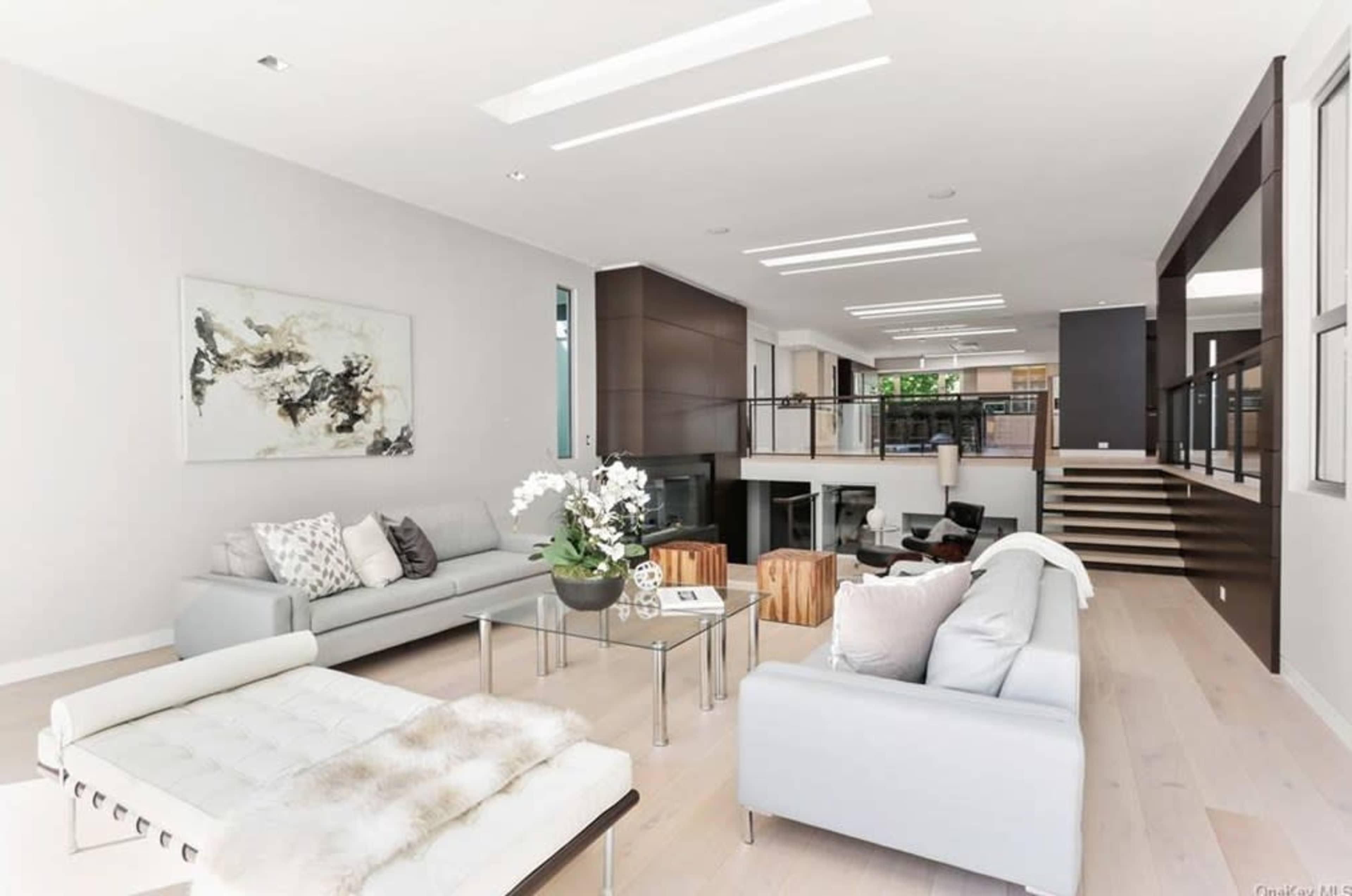 The image shows a spacious modern living room featuring light-colored walls, contemporary furniture, and an open layout that connects to an upper level with a staircase.