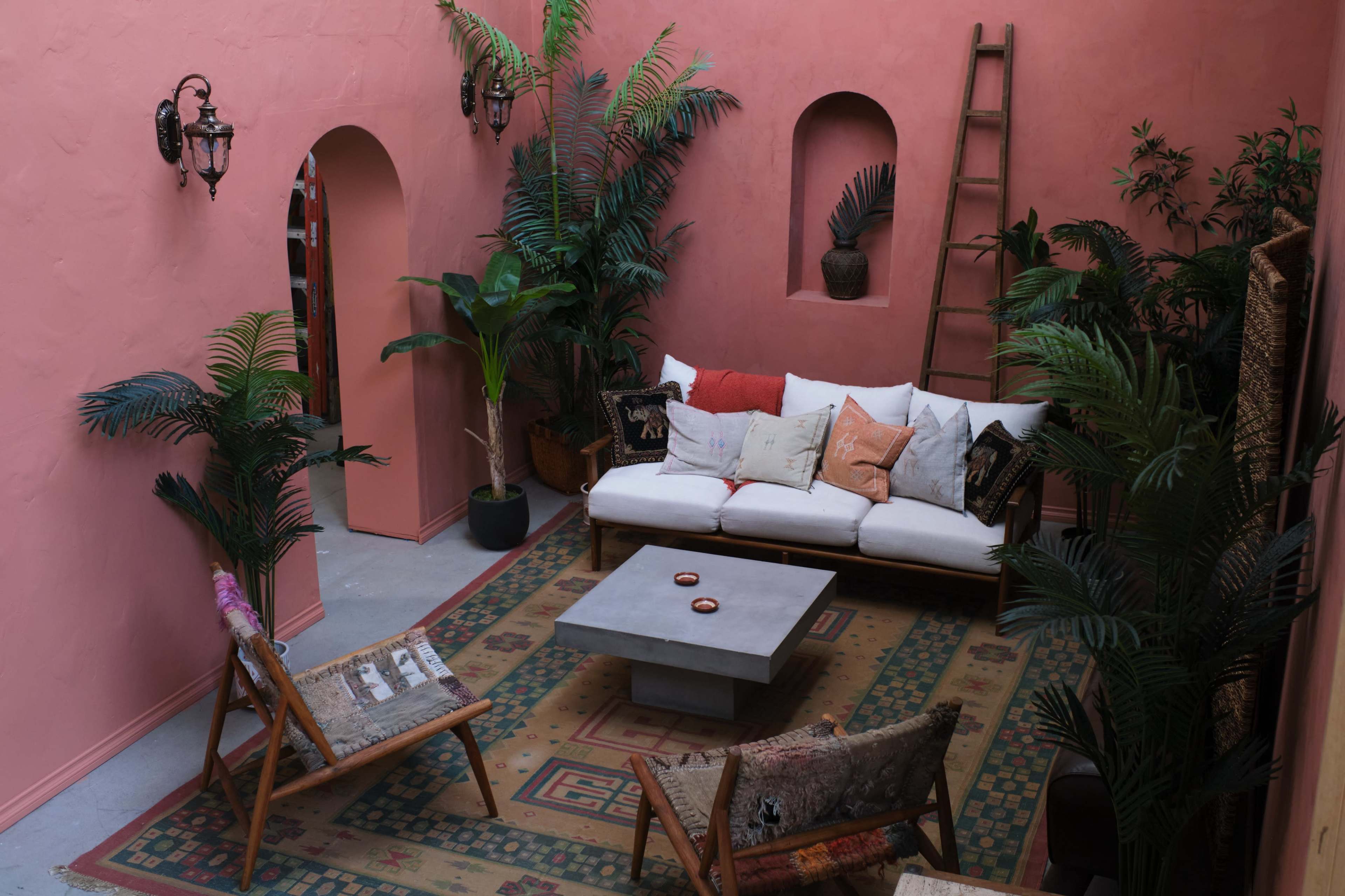A cozy courtyard with pink walls, a white sofa, a concrete coffee table, and greenery from potted plants.