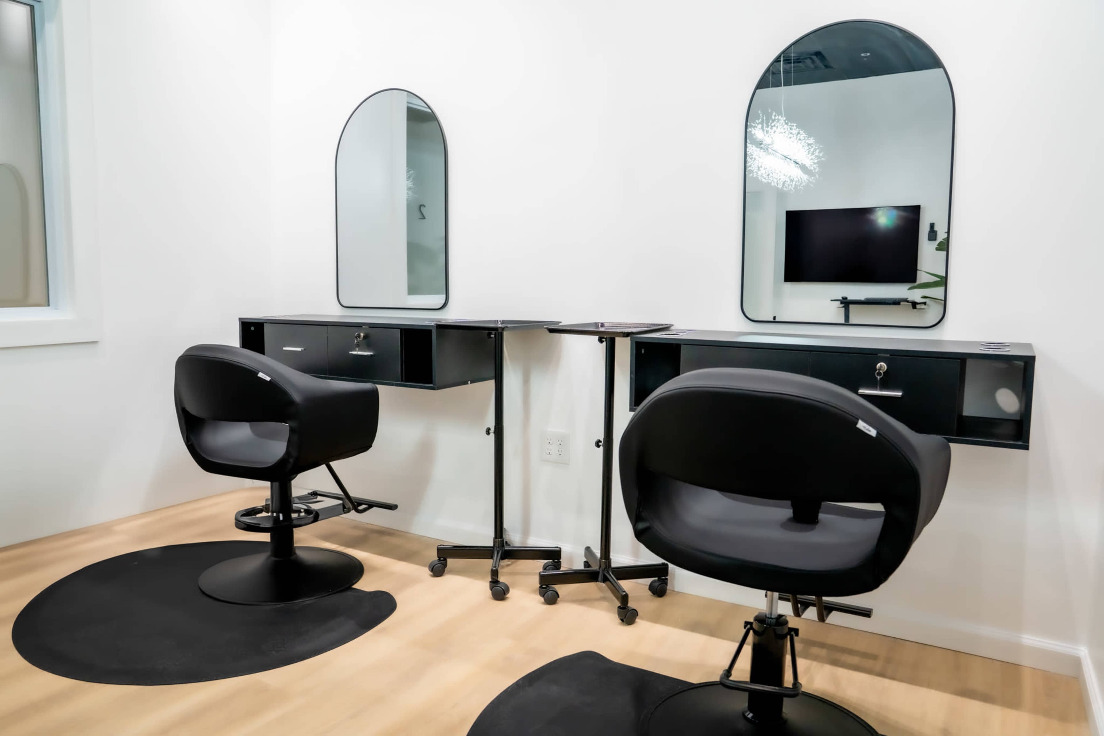 The image shows a modern beauty salon interior with two styling stations featuring mirrors, black chairs, and a mobile table.