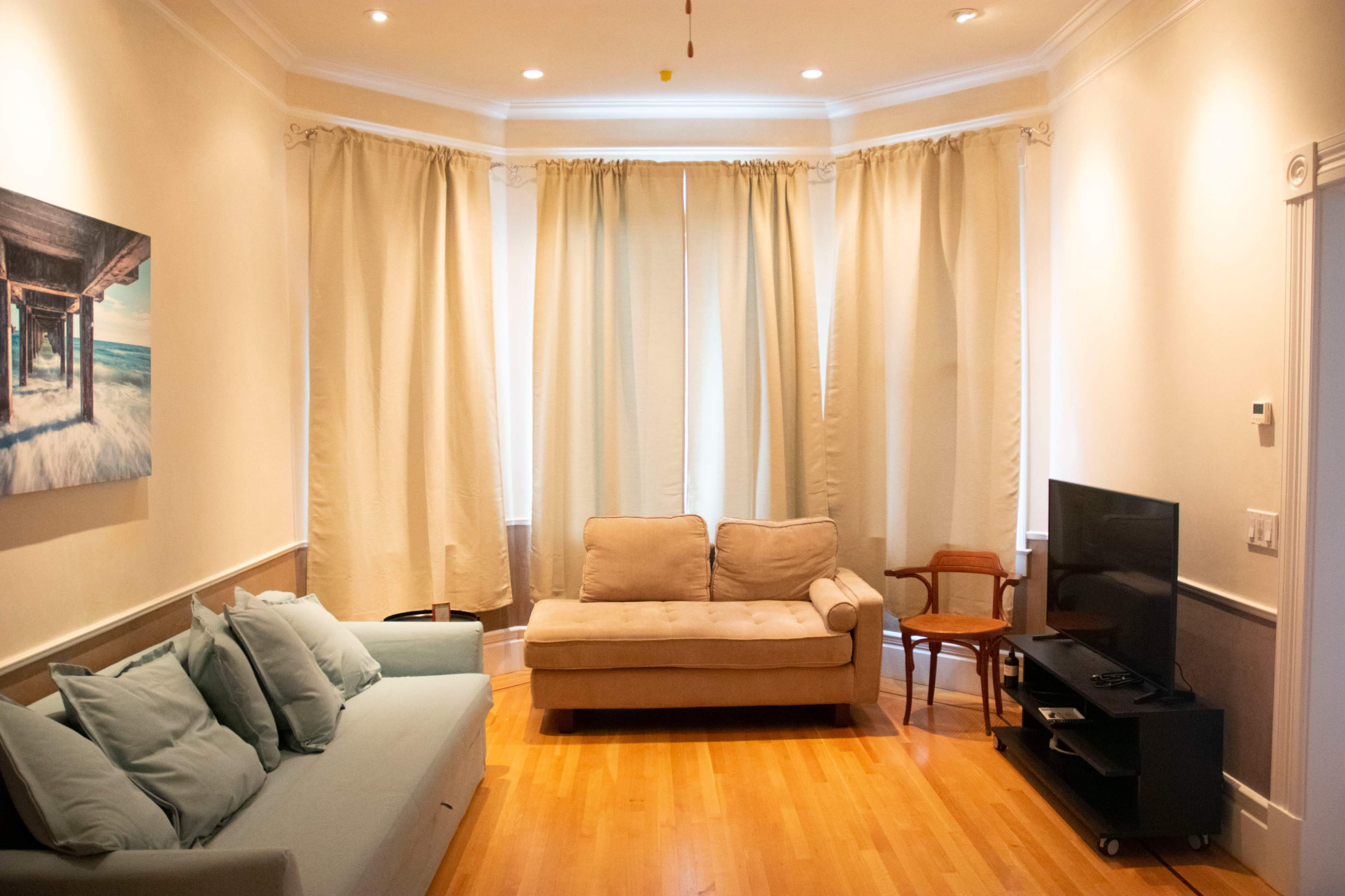 A living room with light wooden floors, a beige sofa, a blue couch, a wooden chair, and a television on a black stand, surrounded by light-colored curtains.