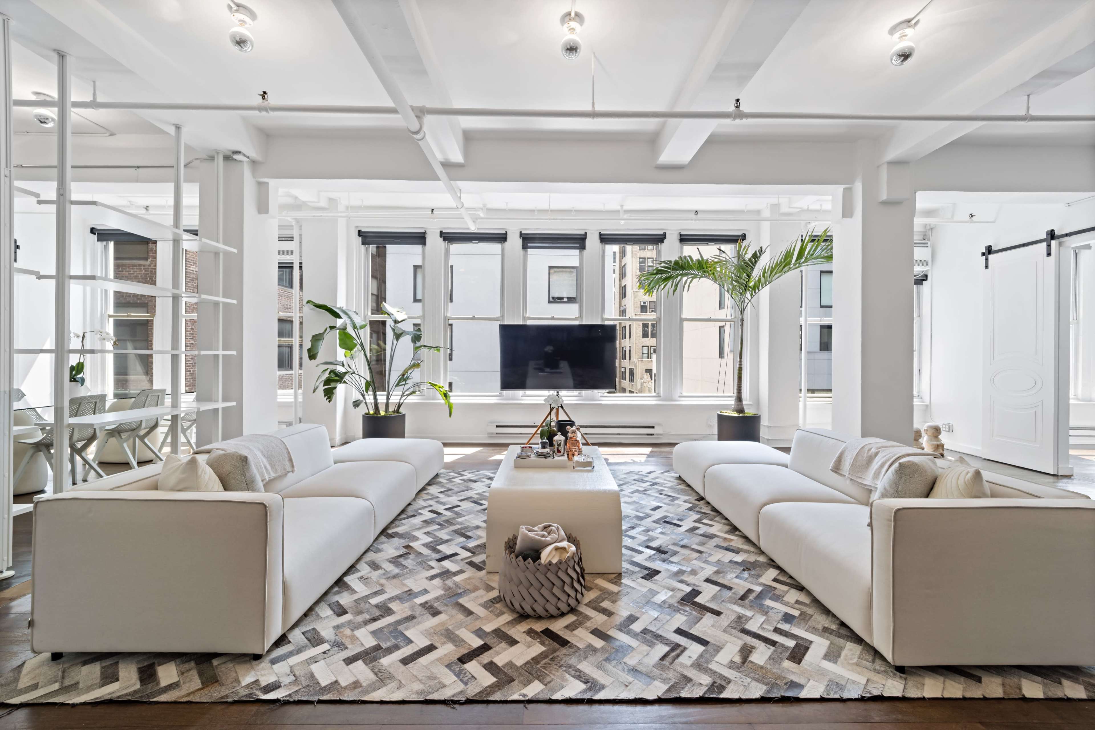A bright, open living room features two white sofas facing each other across a coffee table, with large windows allowing natural light and a plant in the corner.