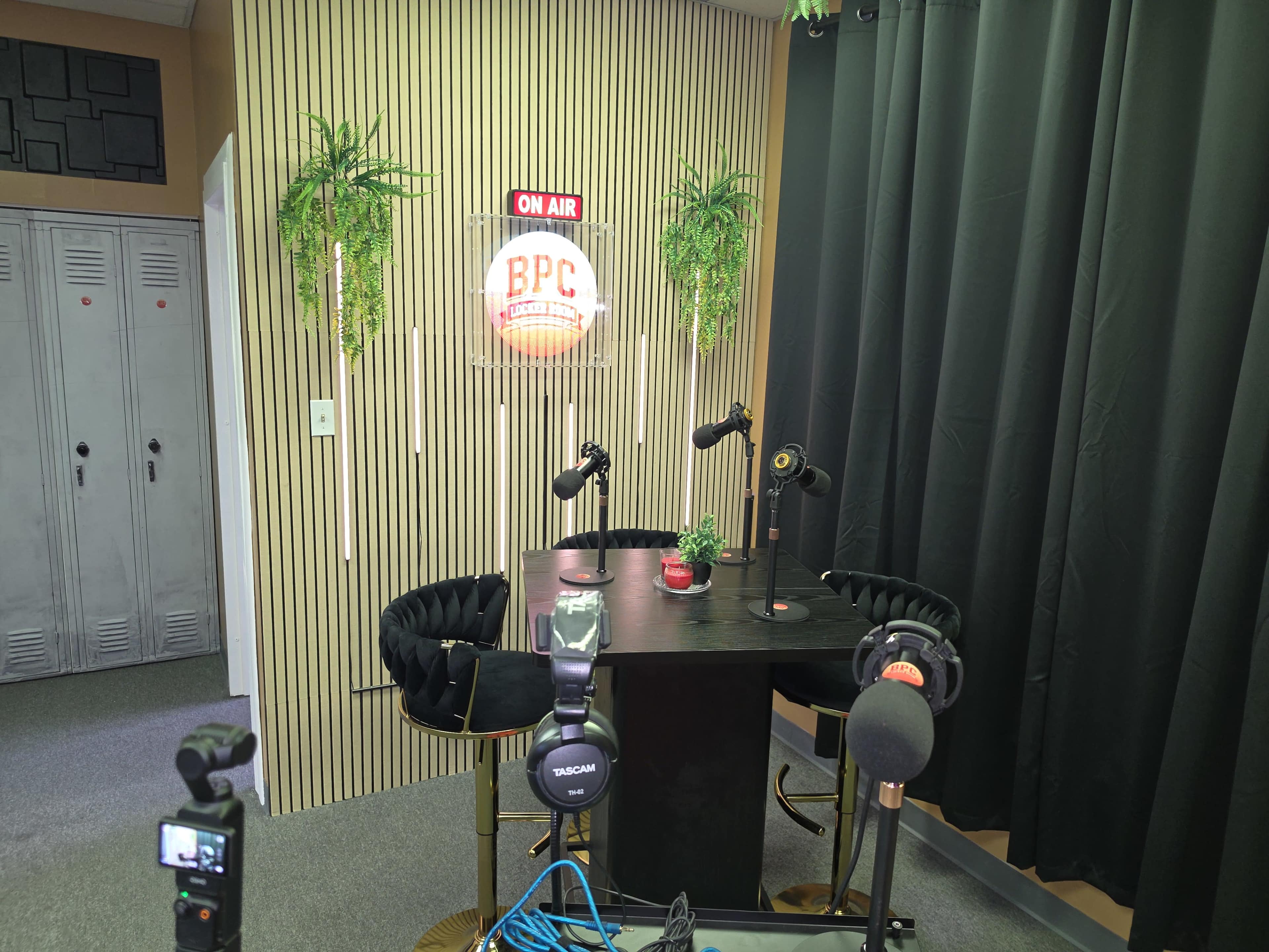 The image shows a podcast recording studio with a round table, two black chairs, microphones, and a neon "ON AIR" sign on the wall.