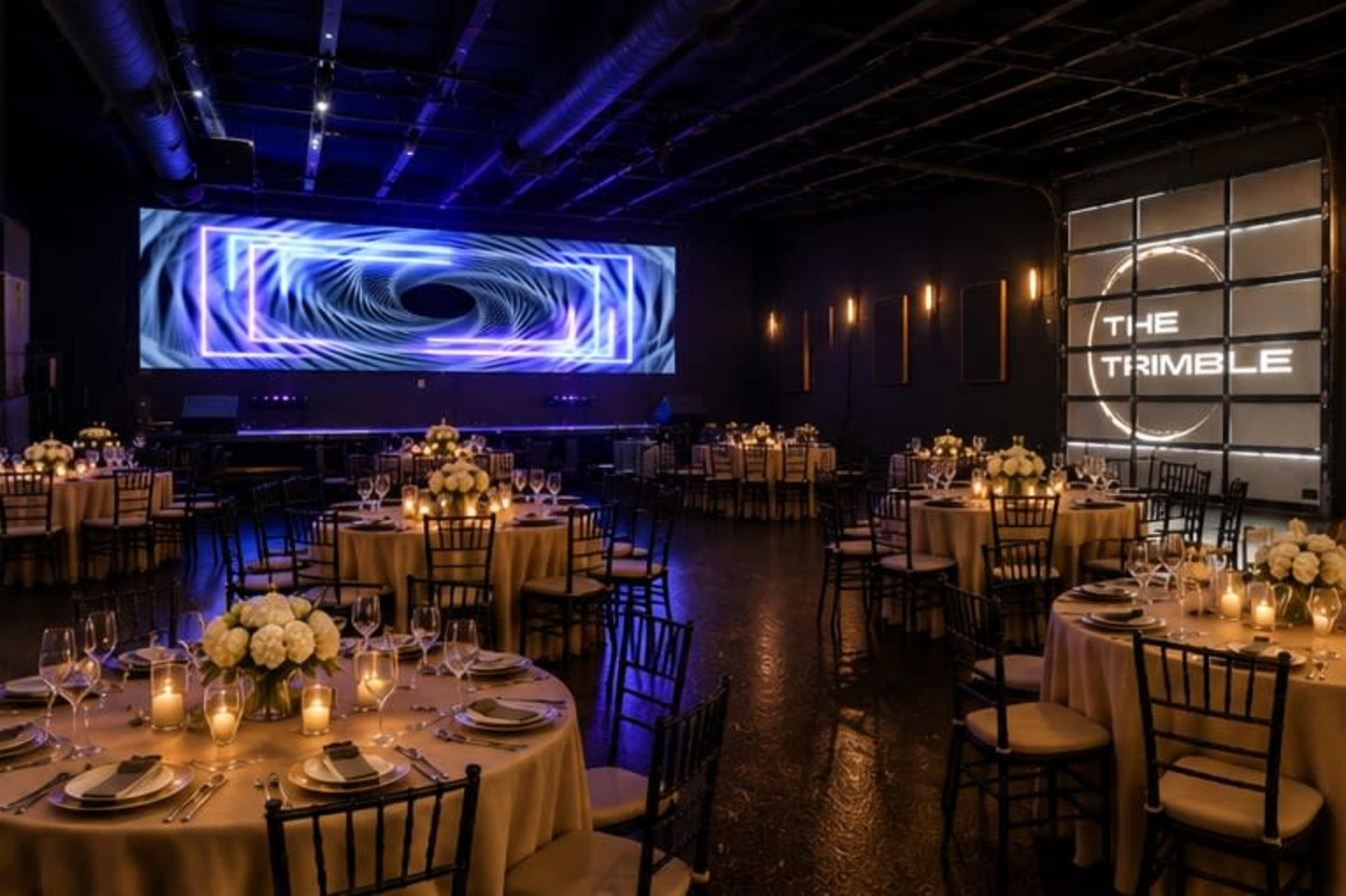 The image shows a modern event space set for a formal dinner, with round tables adorned with flowers and candles, and a large screen displaying abstract visuals in the background.