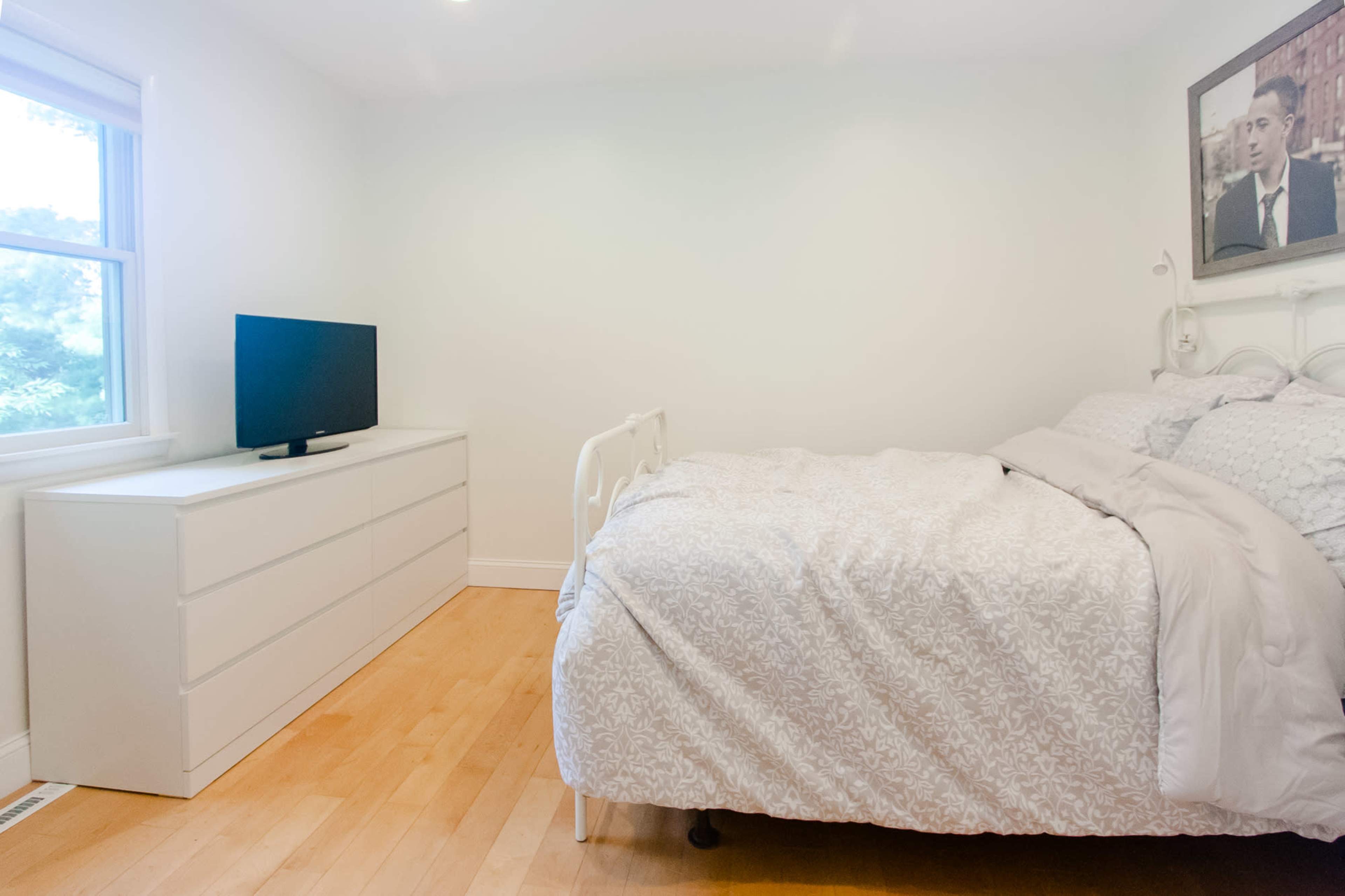 A bright bedroom features a bed with a light-colored duvet, a white dresser, and a television against a wall with a window.