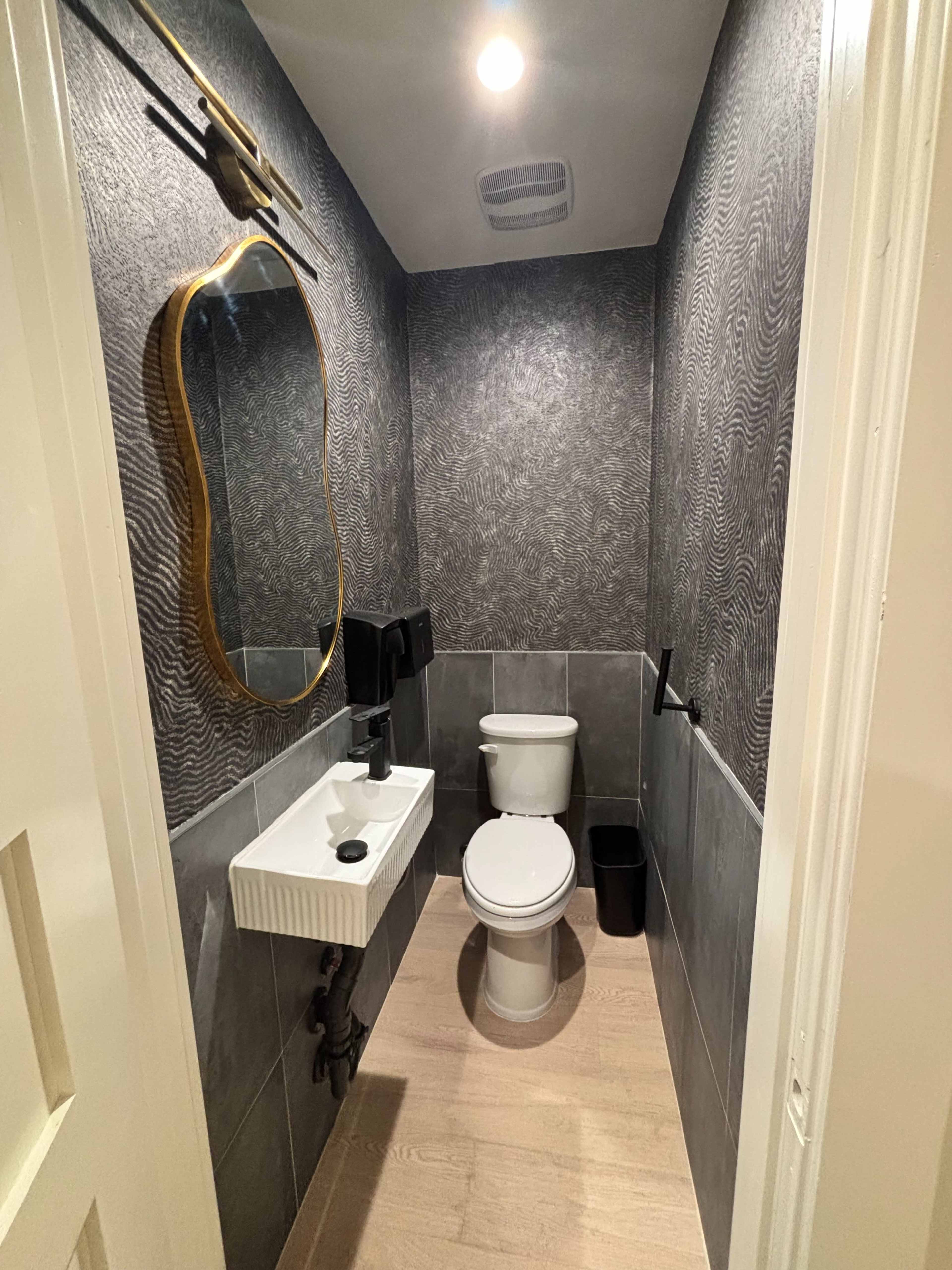 The image shows a small, modern bathroom with textured gray walls, a white toilet, a sleek sink, a round mirror with a gold frame, and a black trash can.