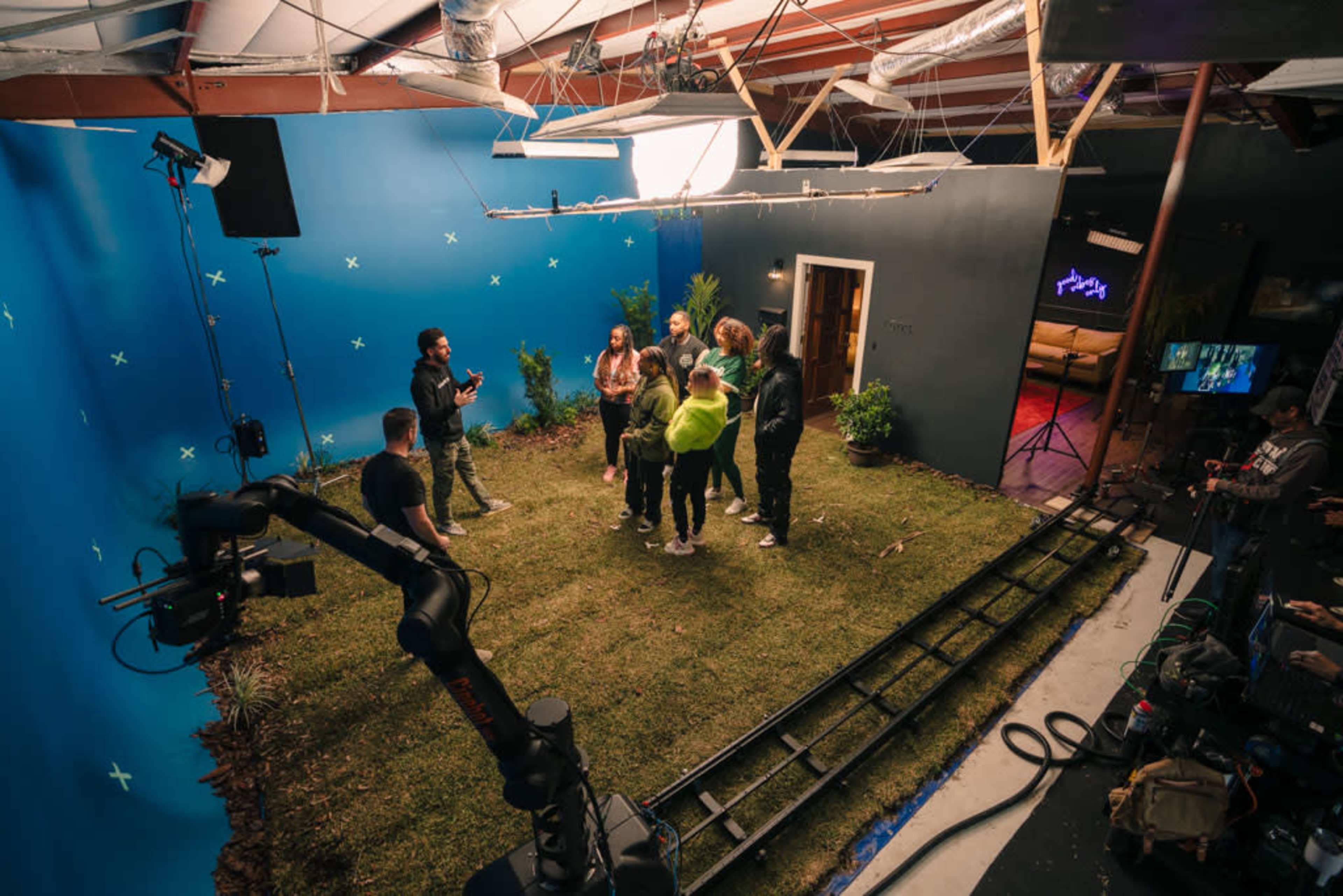 A group of people stands on a set with artificial grass, surrounded by plants and a blue backdrop, while a camera and crew are positioned nearby.