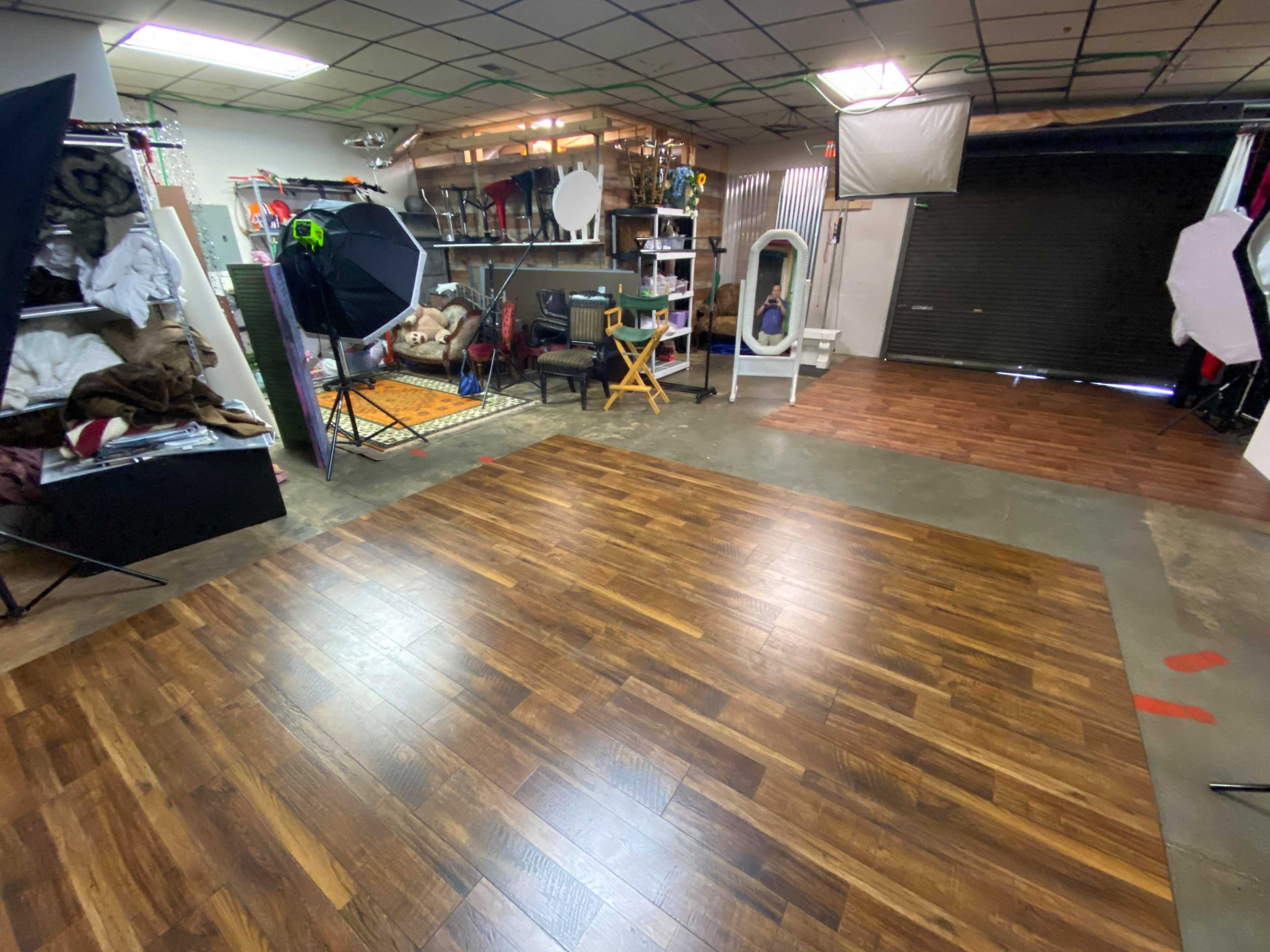 The image shows a photography studio with wooden flooring, equipped with various photo backdrops, lighting equipment, and furniture arranged in distinct areas.