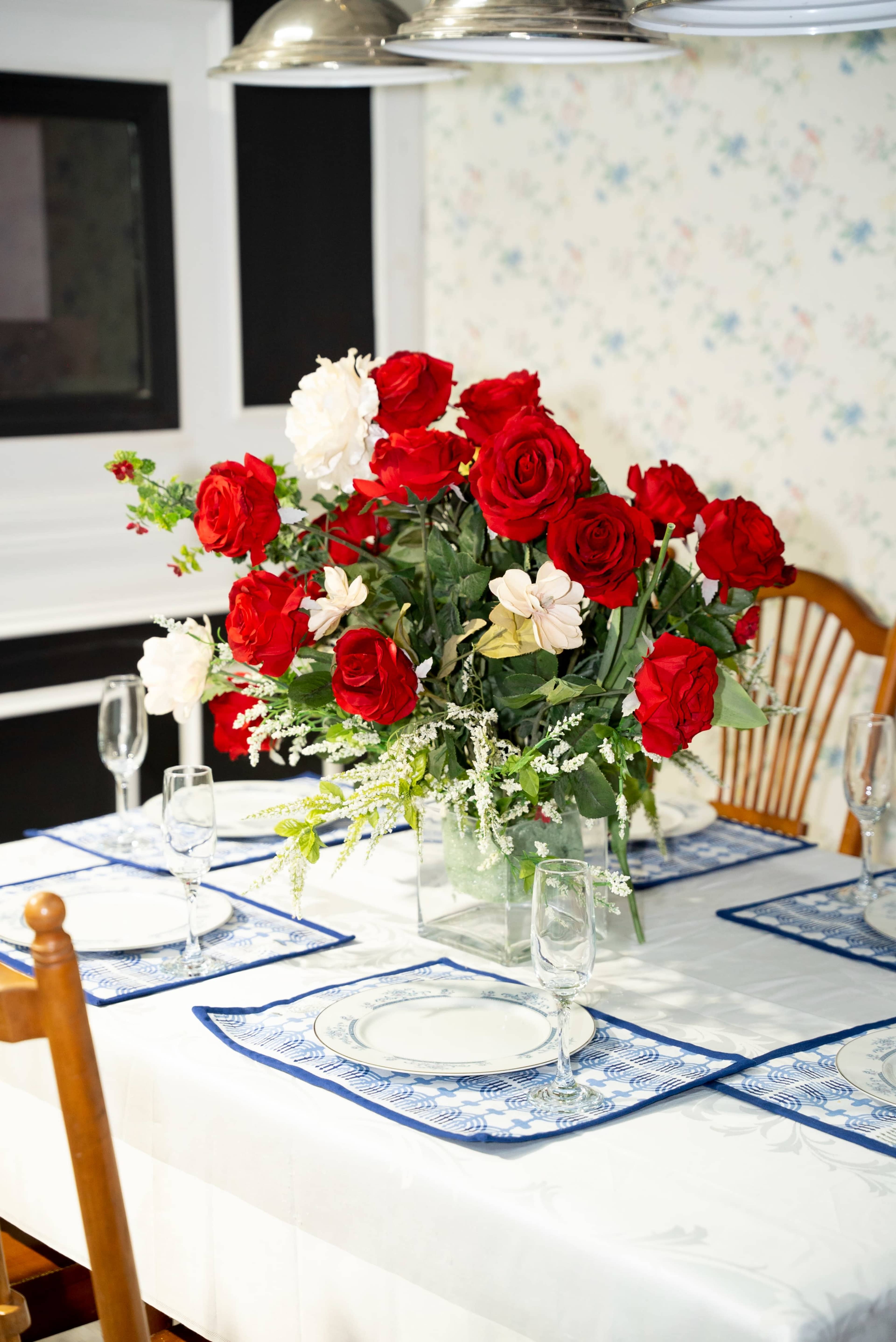 A dining table is set with plates and glasses, featuring a large vase filled with red roses and white flowers as a centerpiece.