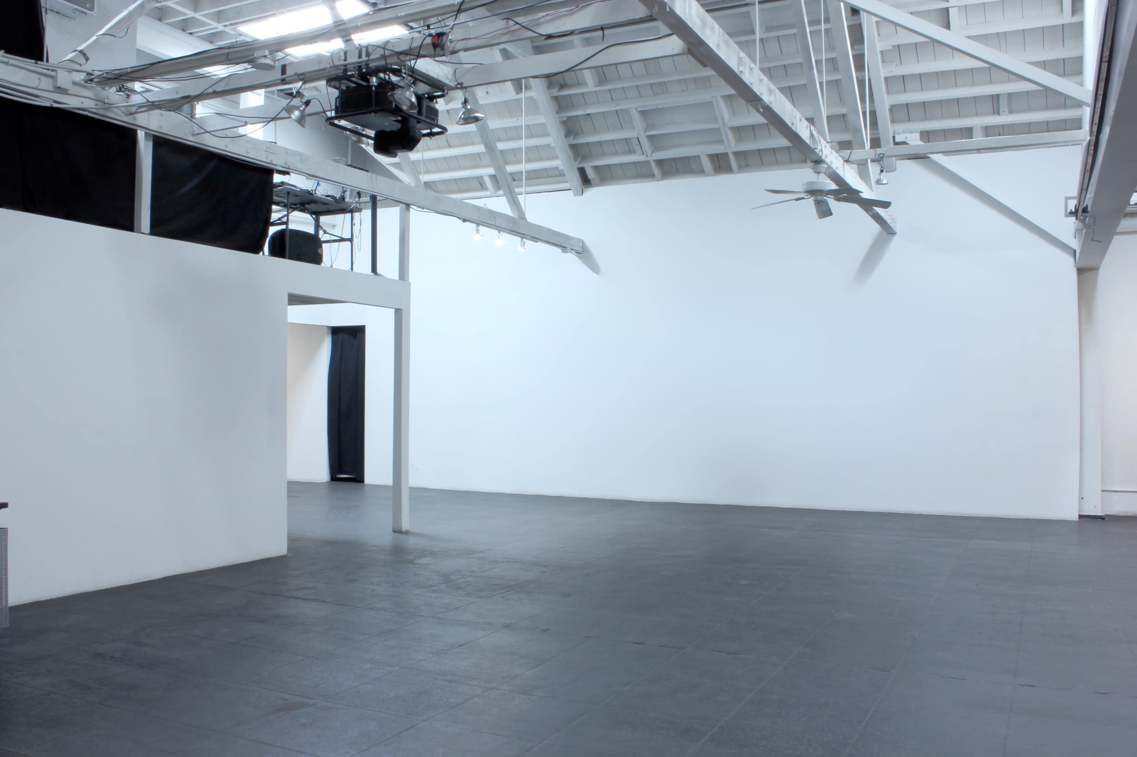 The image shows a spacious, empty gallery with high ceilings, large white walls, and a dark tiled floor.