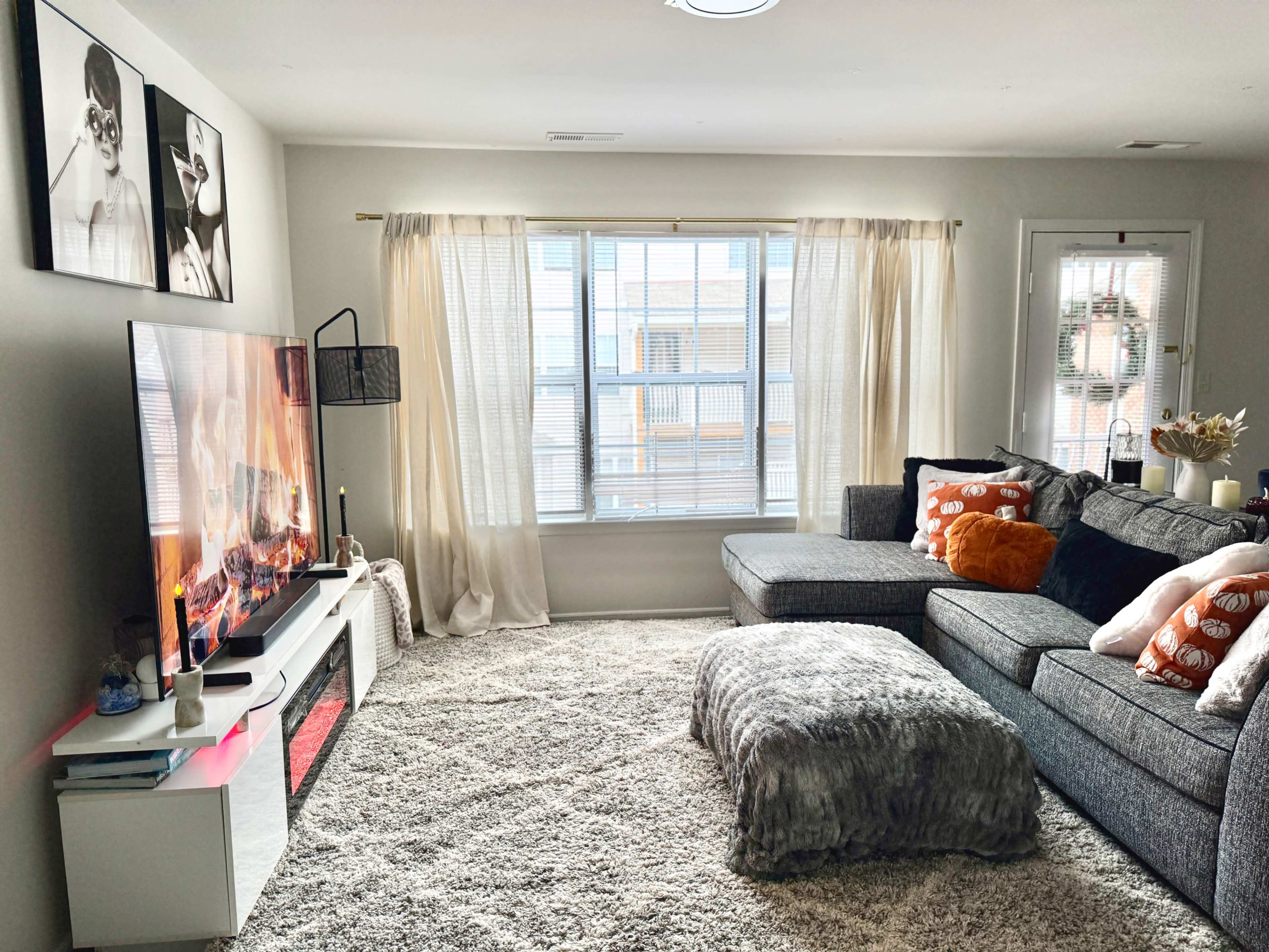 The image shows a cozy living room with a large sectional sofa, a television displaying a fireplace, and a window with sheer curtains letting in natural light.