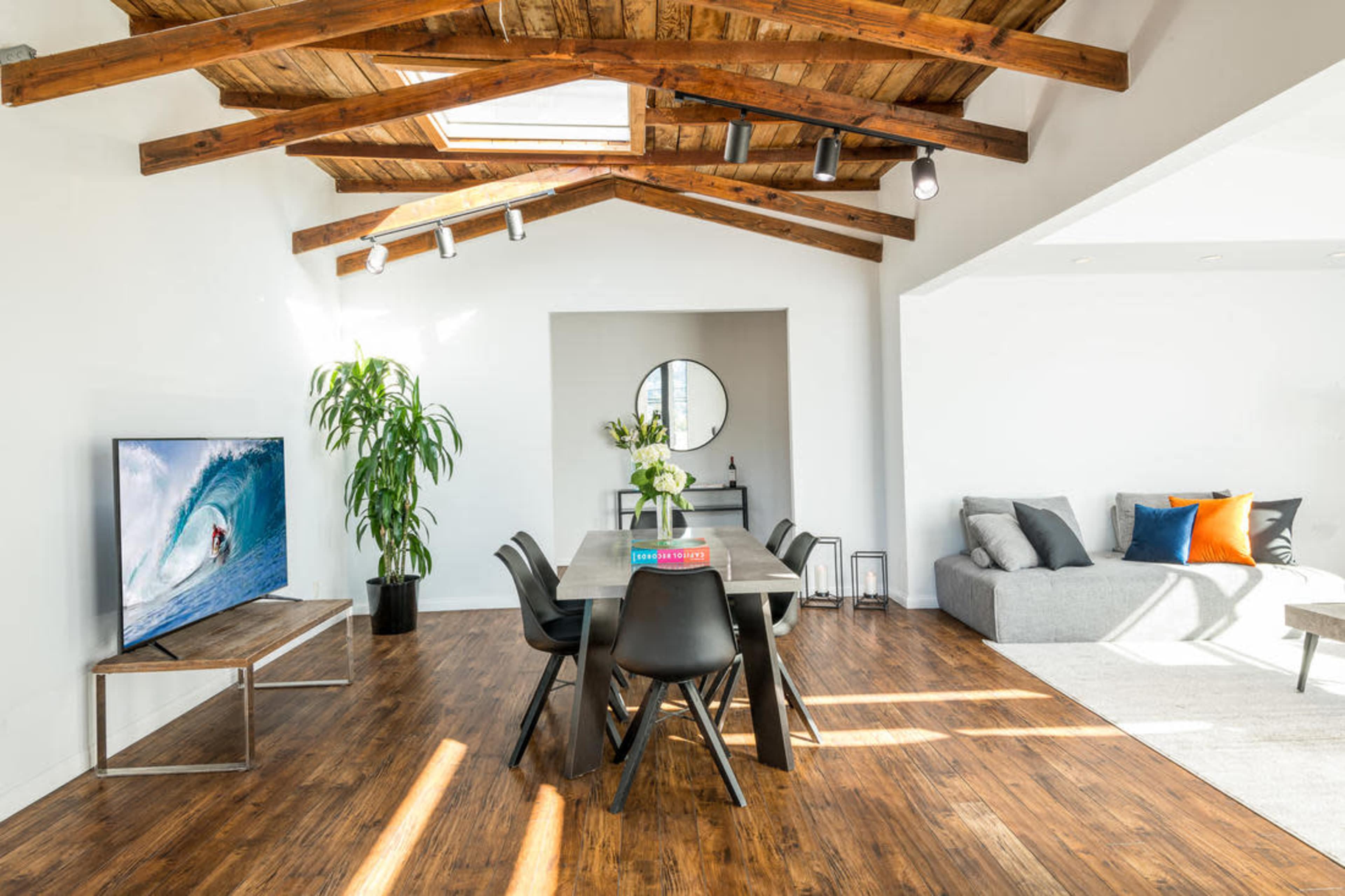 A modern living space features a dining table with black chairs, a large television, a cozy gray sofa, and exposed wooden beams on the ceiling.