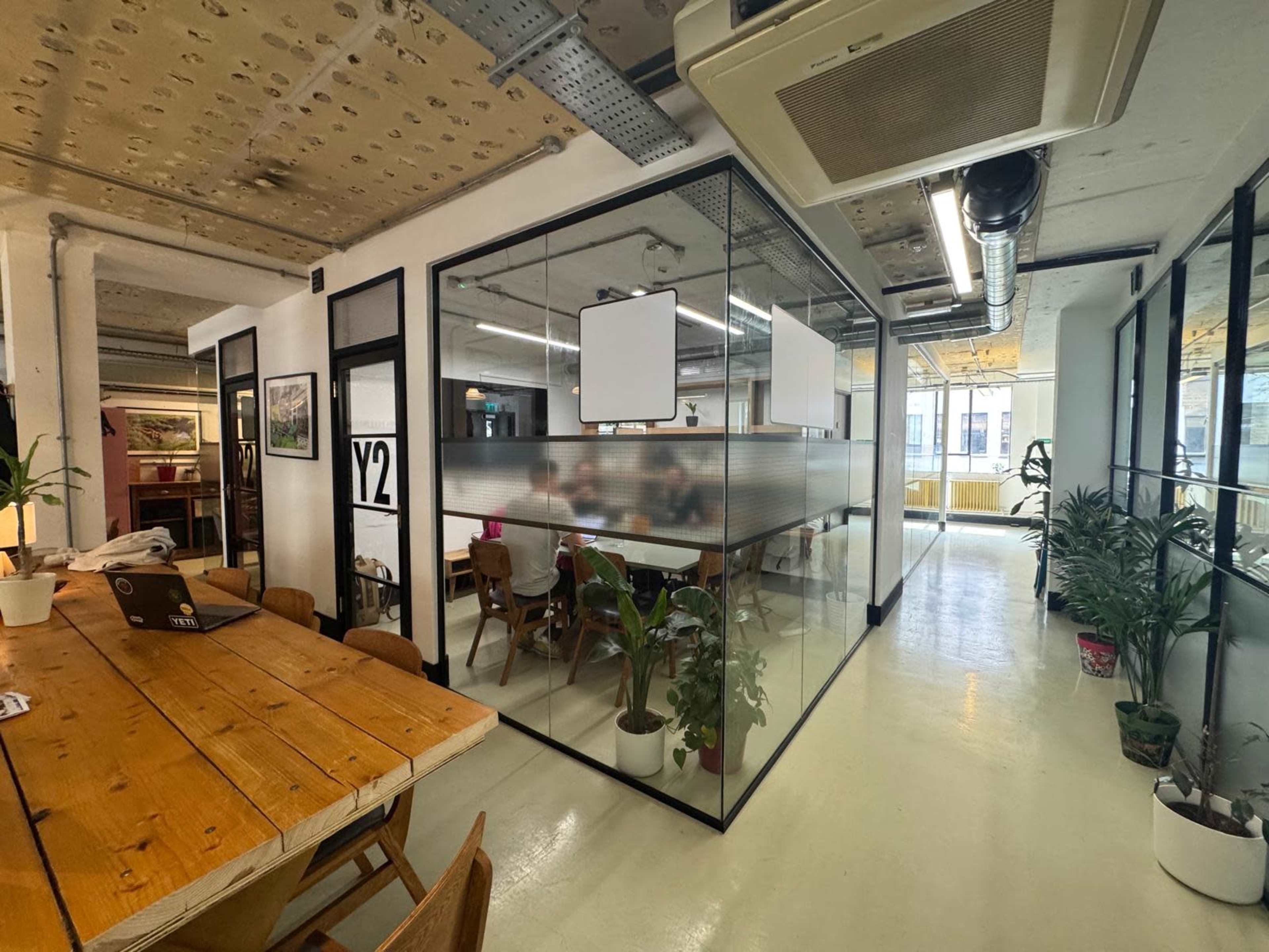 The image shows an open office space with glass-walled meeting rooms, a large wooden table, and several plants throughout.