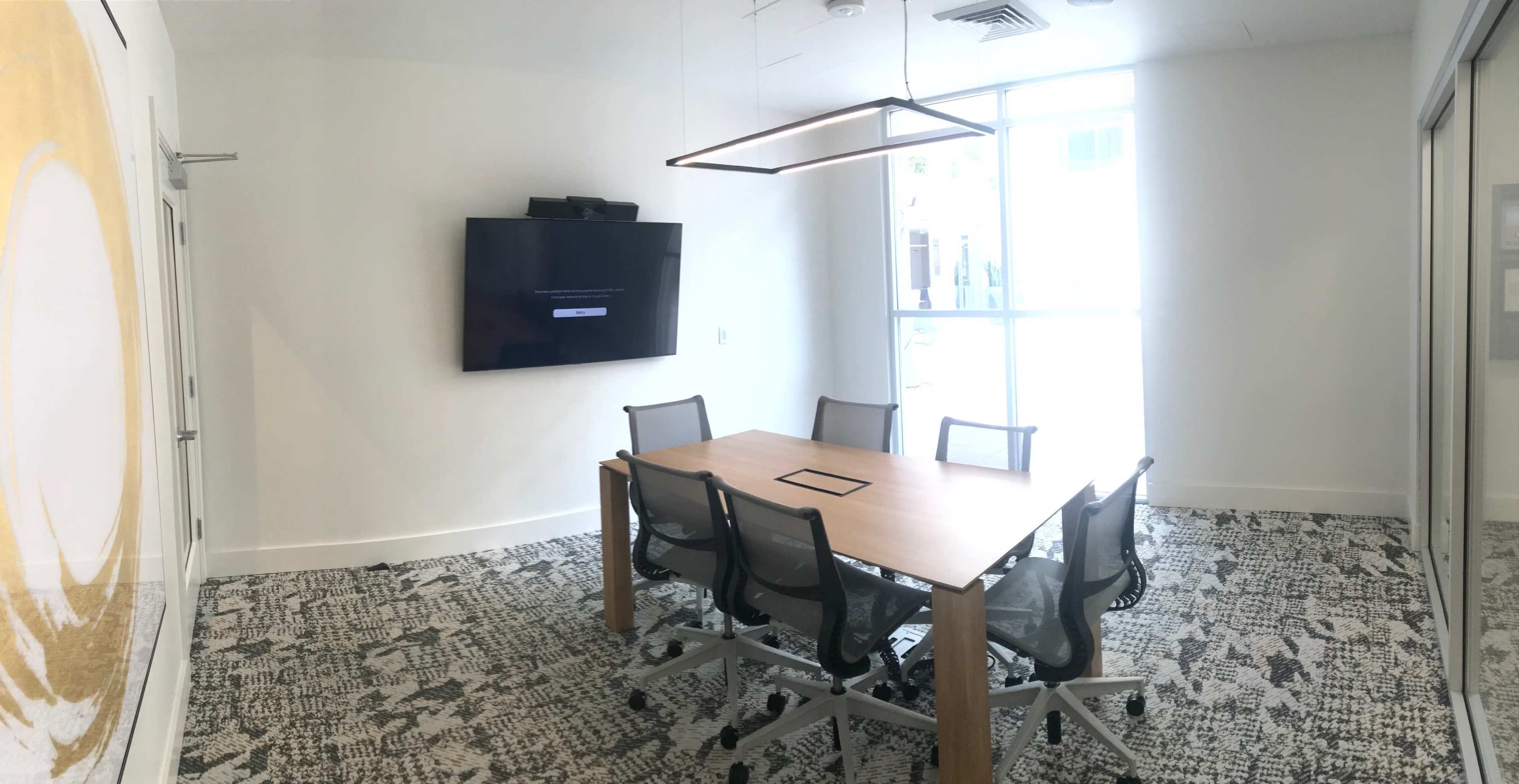 A modern conference room features a rectangular wooden table surrounded by six chairs, a large wall-mounted screen, and ample natural light from a nearby window.
