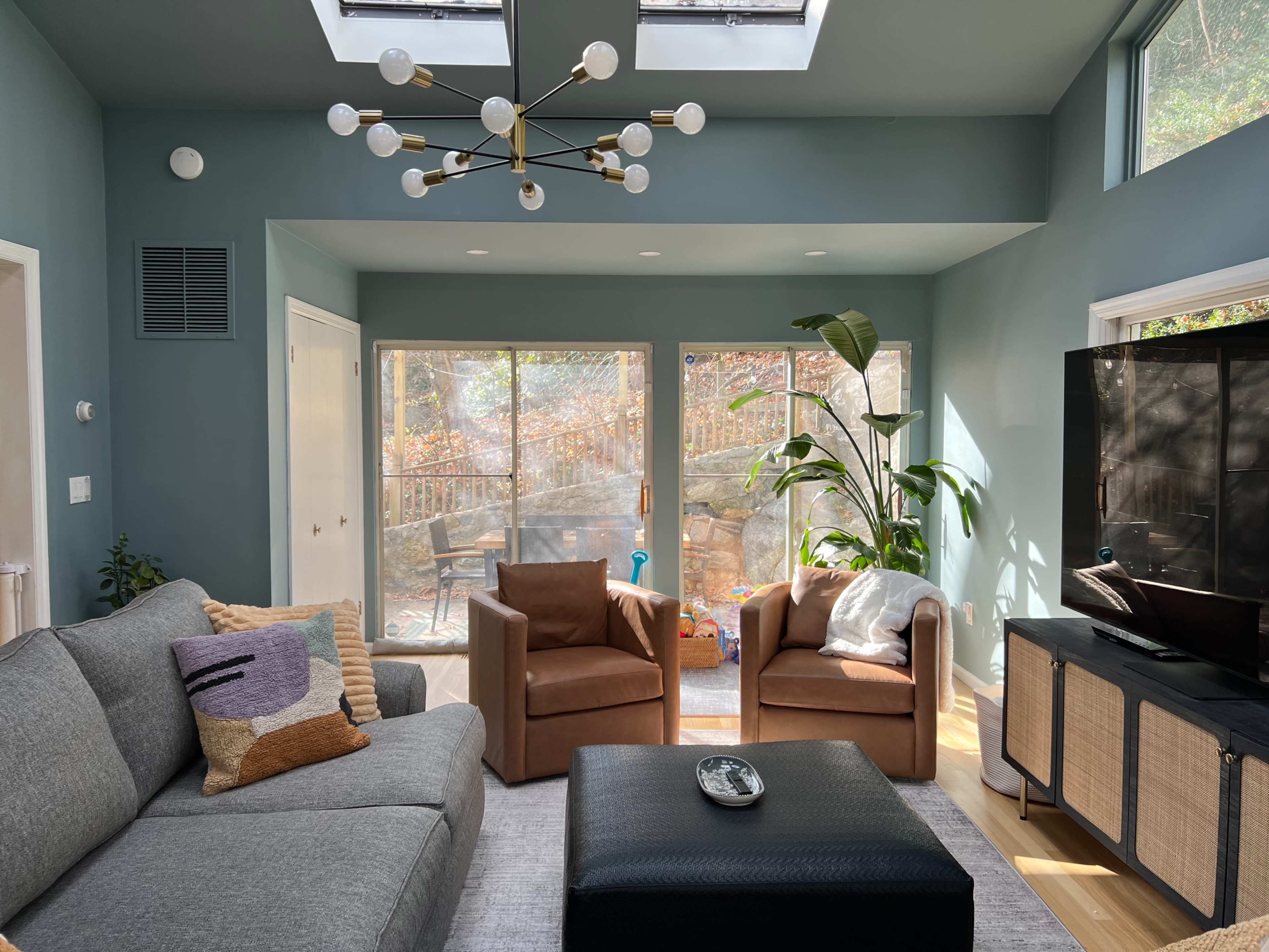 A living room with a gray sofa, two brown armchairs, large windows overlooking an outdoor space, and a decorative light fixture on the ceiling.