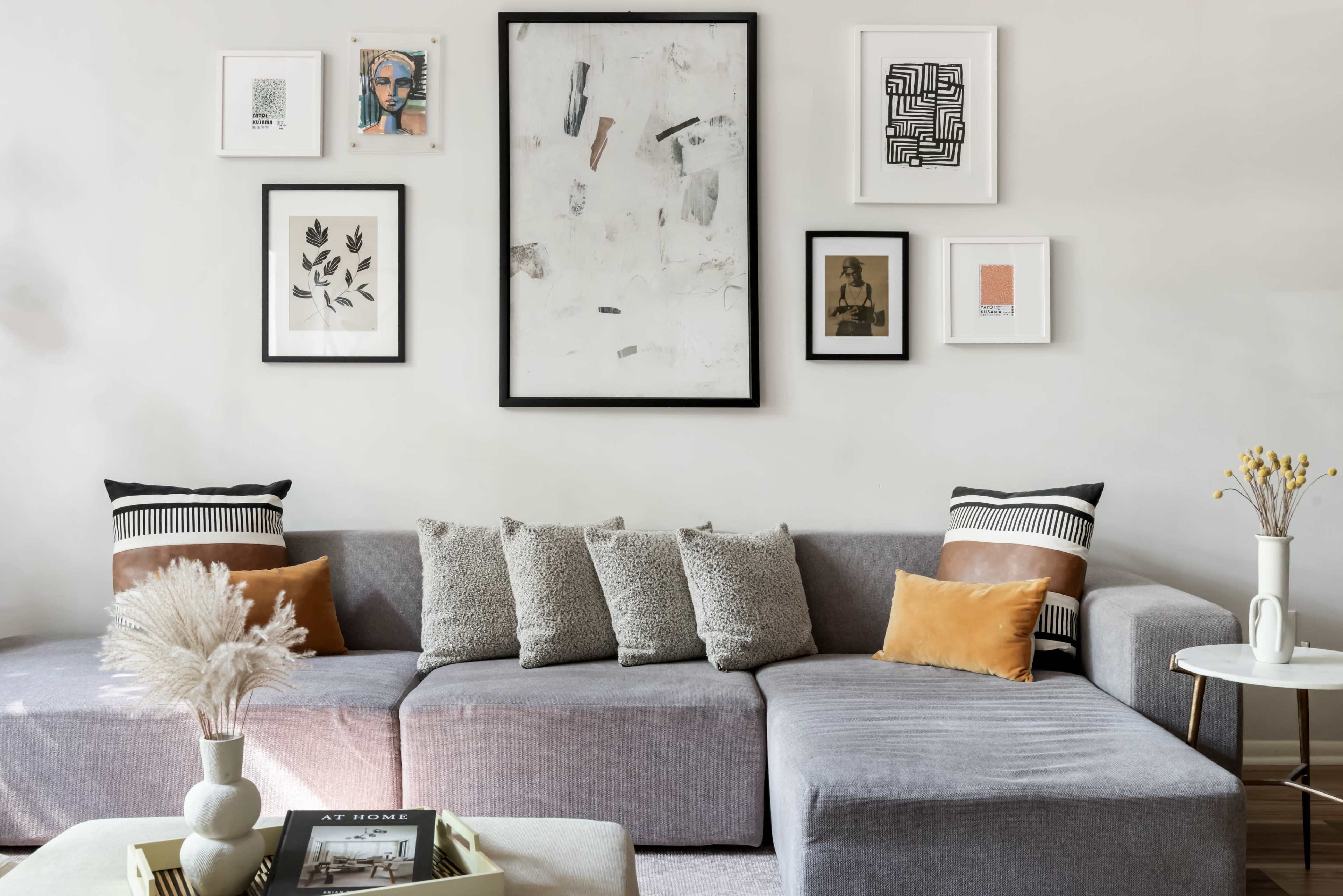 The image shows a modern living room with a gray sectional sofa adorned with decorative pillows, and various framed artworks arranged on the wall above.