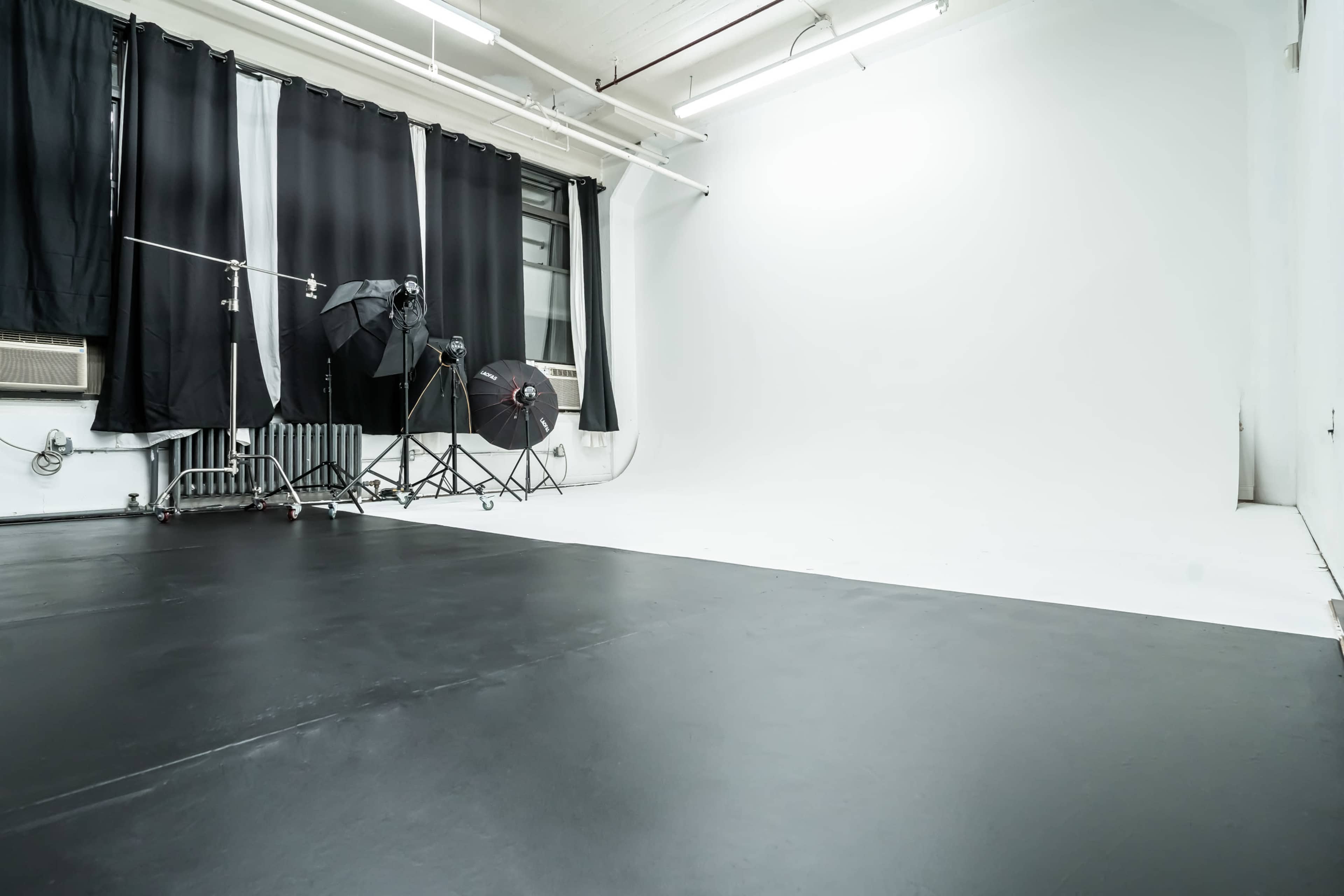 The image shows an empty photography studio with black flooring, backdrop curtains, and lighting equipment positioned in the corner.
