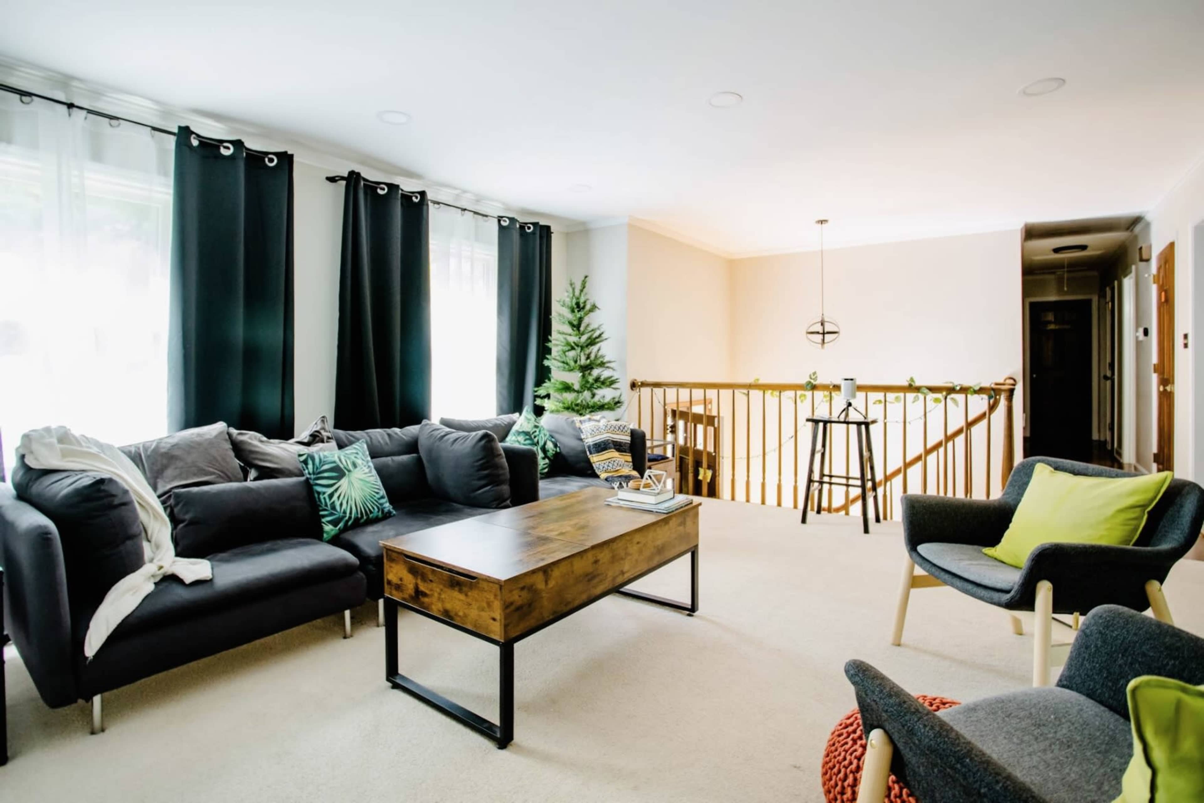 The living room features a dark sectional sofa with decorative cushions, a wooden coffee table, a small Christmas tree, and a staircase in the background.