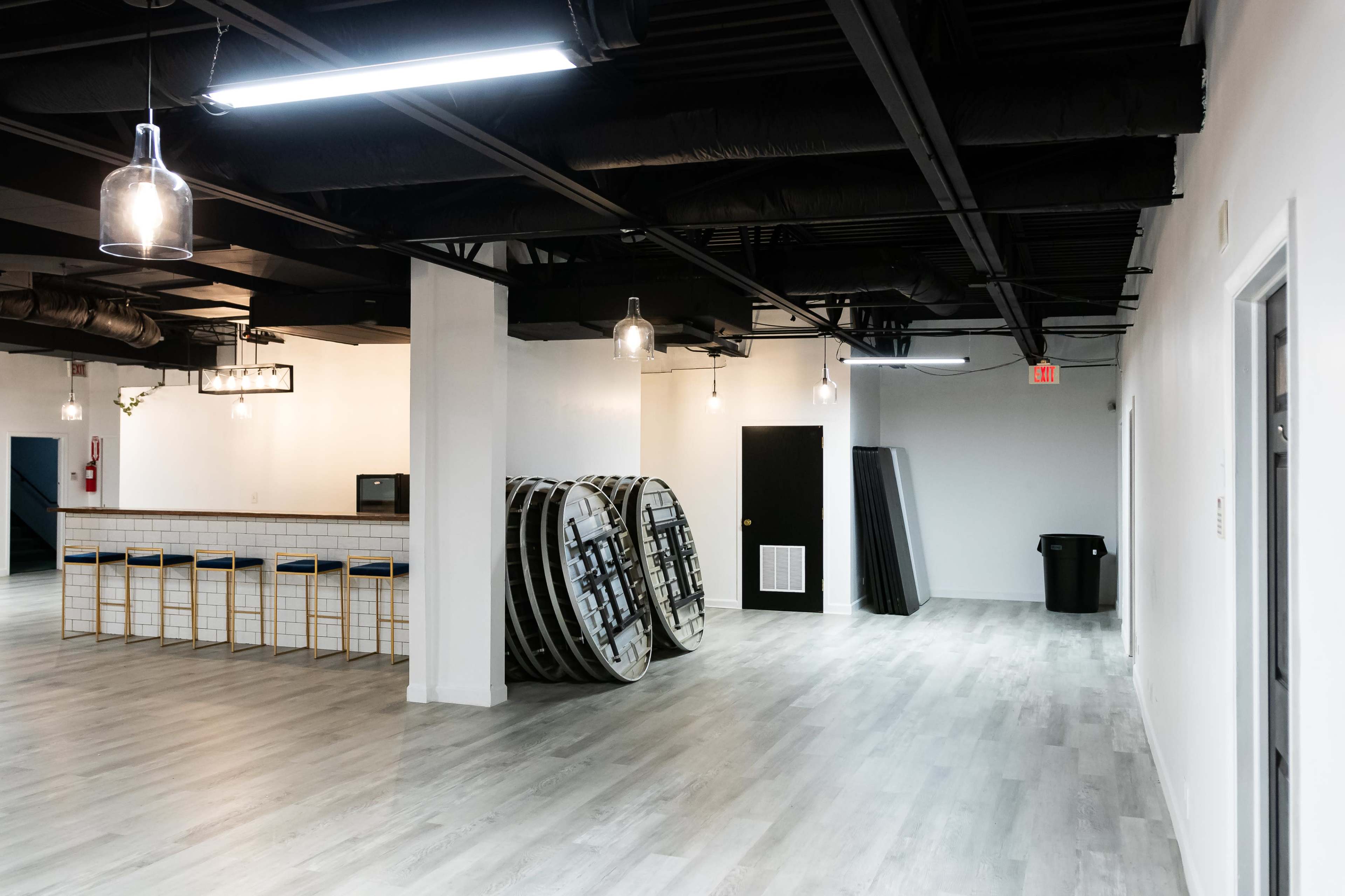 The image shows a spacious, well-lit room with a bar area, stacked round tables, and a black trash can near a door in the background.