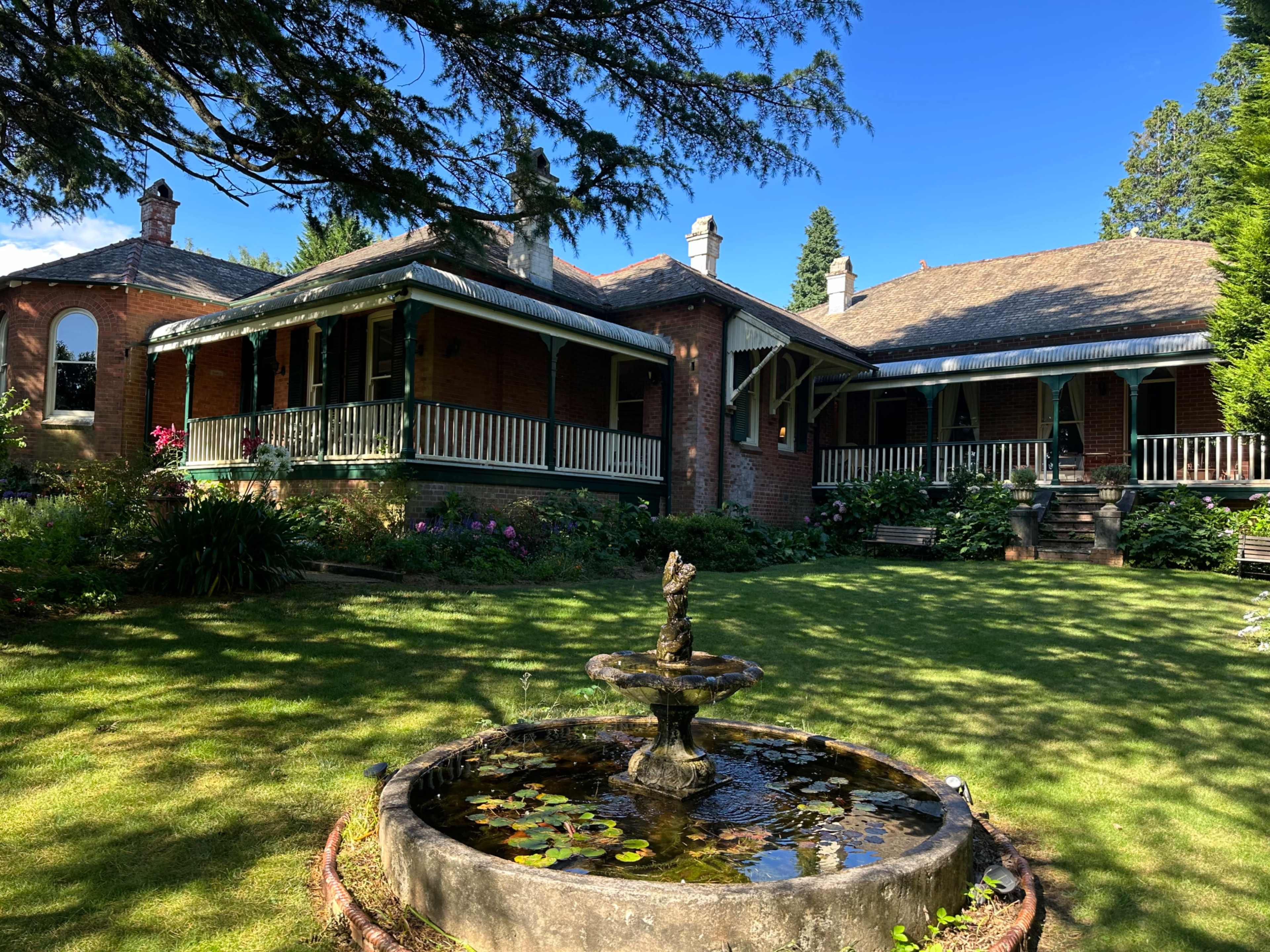 The image shows a large brick house with a surrounding garden featuring a central fountain and well-maintained lawn.