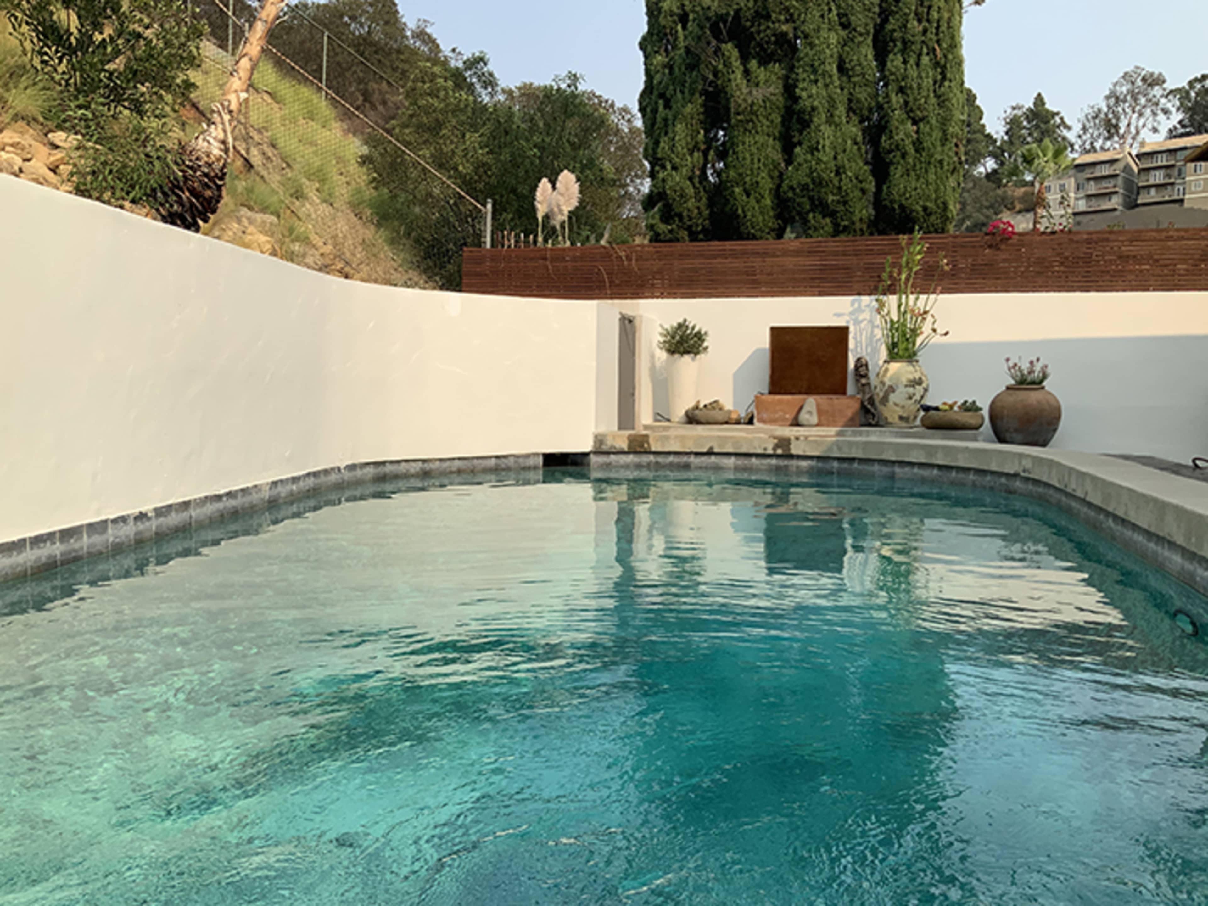 The image shows a clear swimming pool surrounded by a white wall, with greenery and decorative elements in the background.