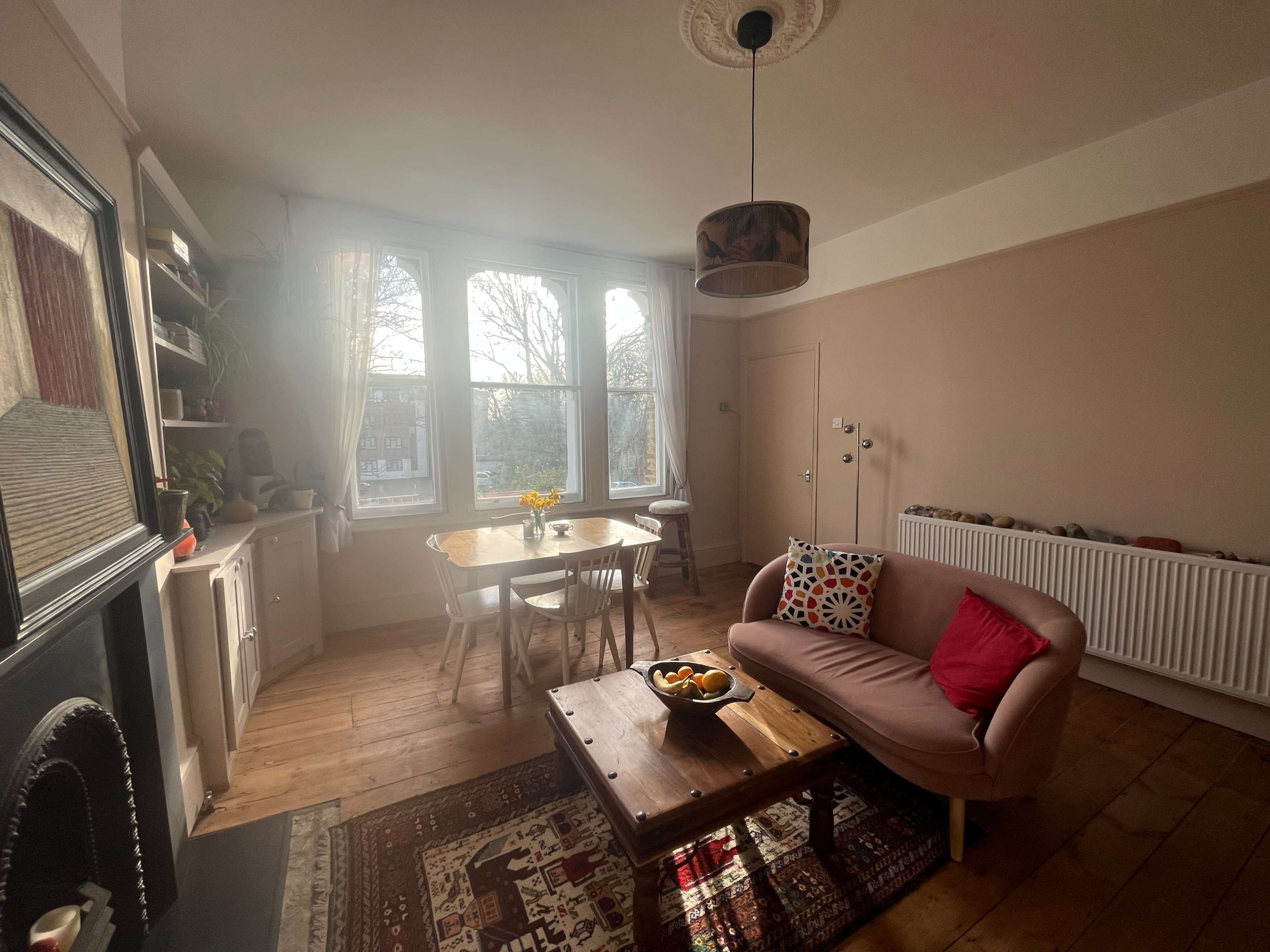 A cozy living room features a wooden floor, a round table with chairs, a sofa adorned with cushions, and large windows letting in natural light.