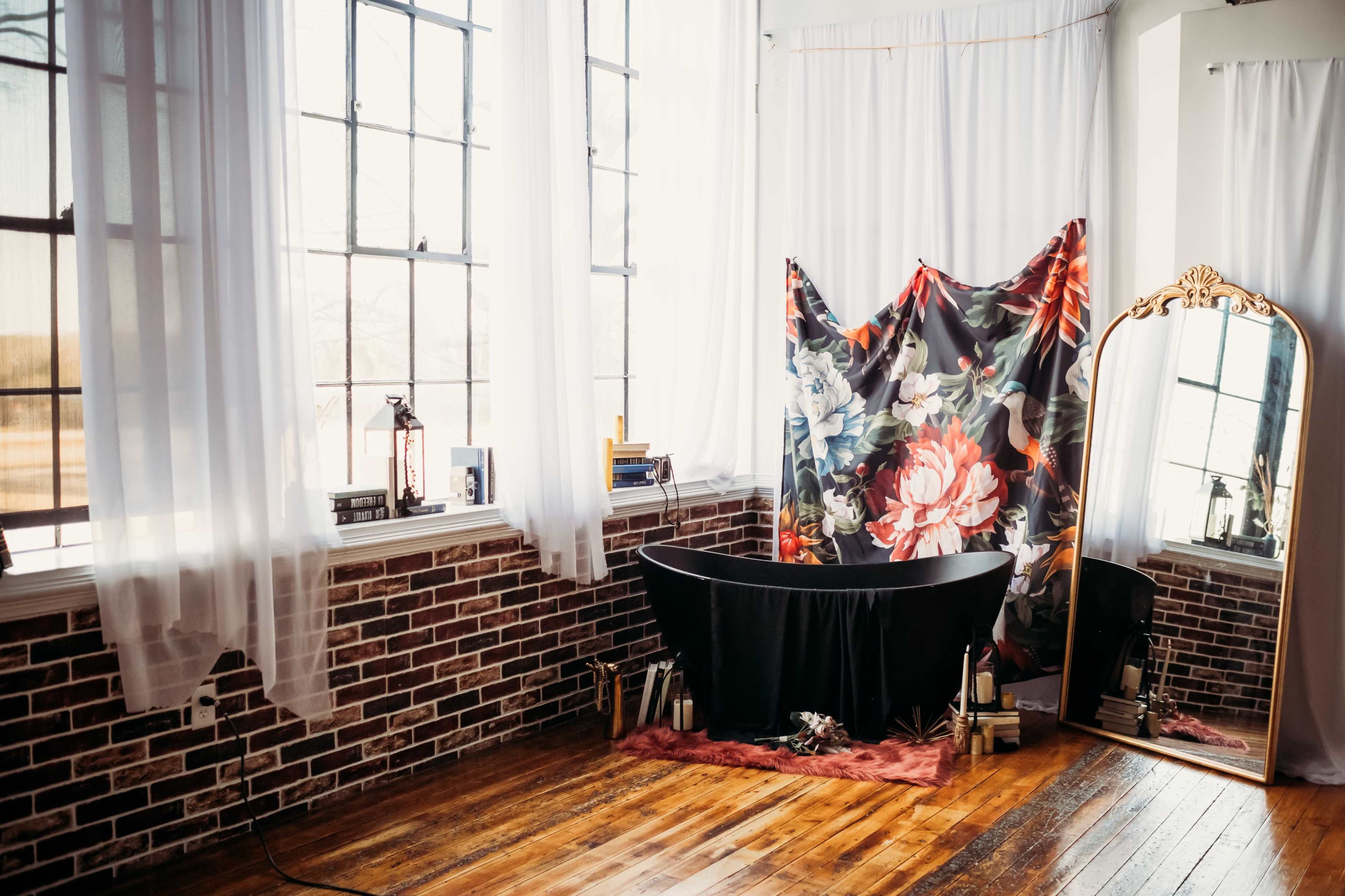 A minimalistic room with large windows, a freestanding black bathtub, a floral backdrop, a mirror, and wooden flooring.