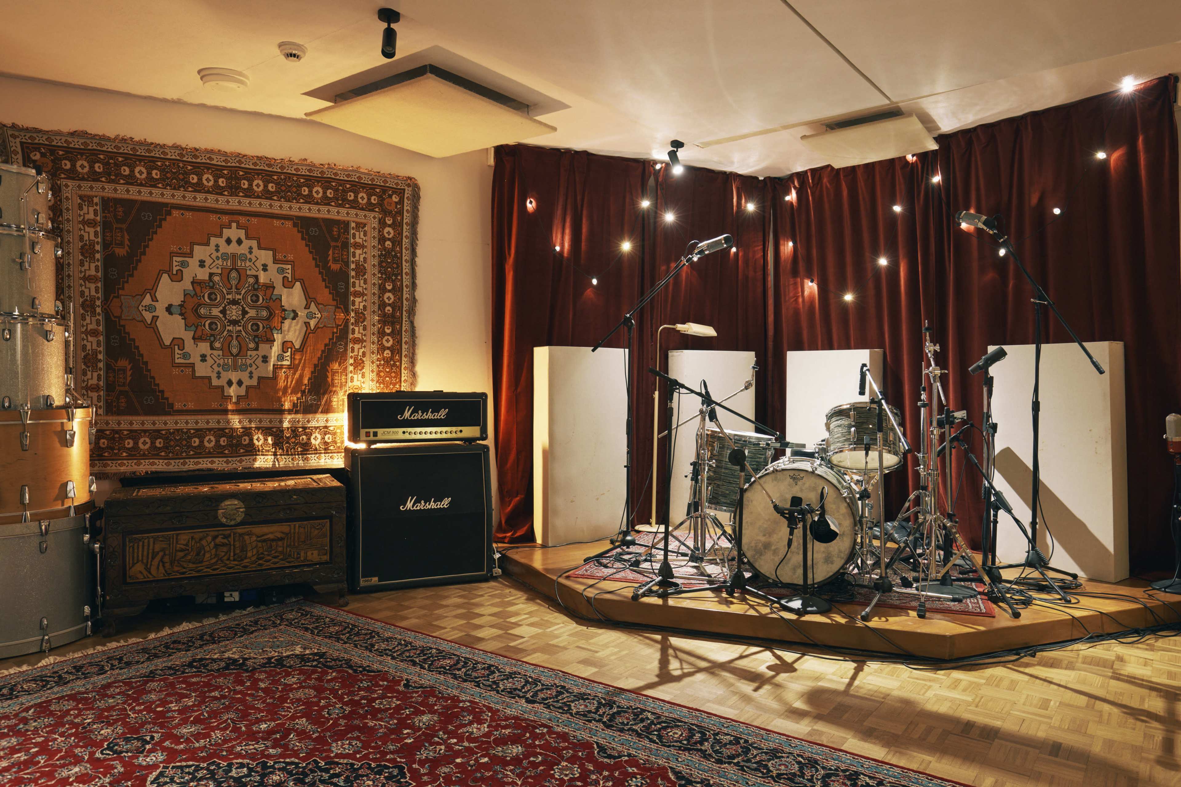 A recording studio is configured with a drum set, amplifiers, and acoustic panels, set against a backdrop of decorative rugs and soft lighting.