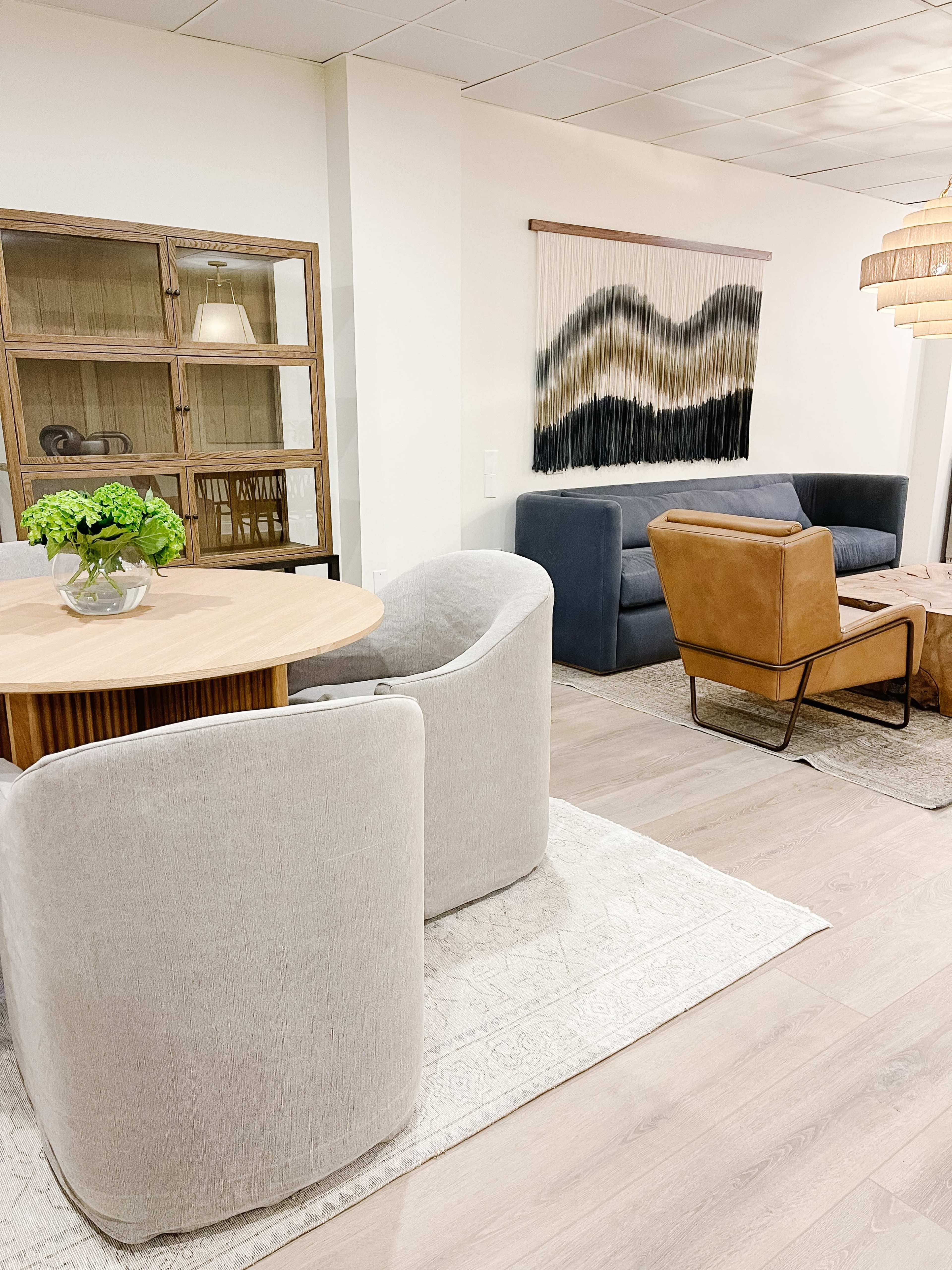A modern living space featuring a round dining table with curved chairs, a wooden display cabinet, a textured wall hanging, and a mix of seating arrangements including a leather armchair and a gray sofa.