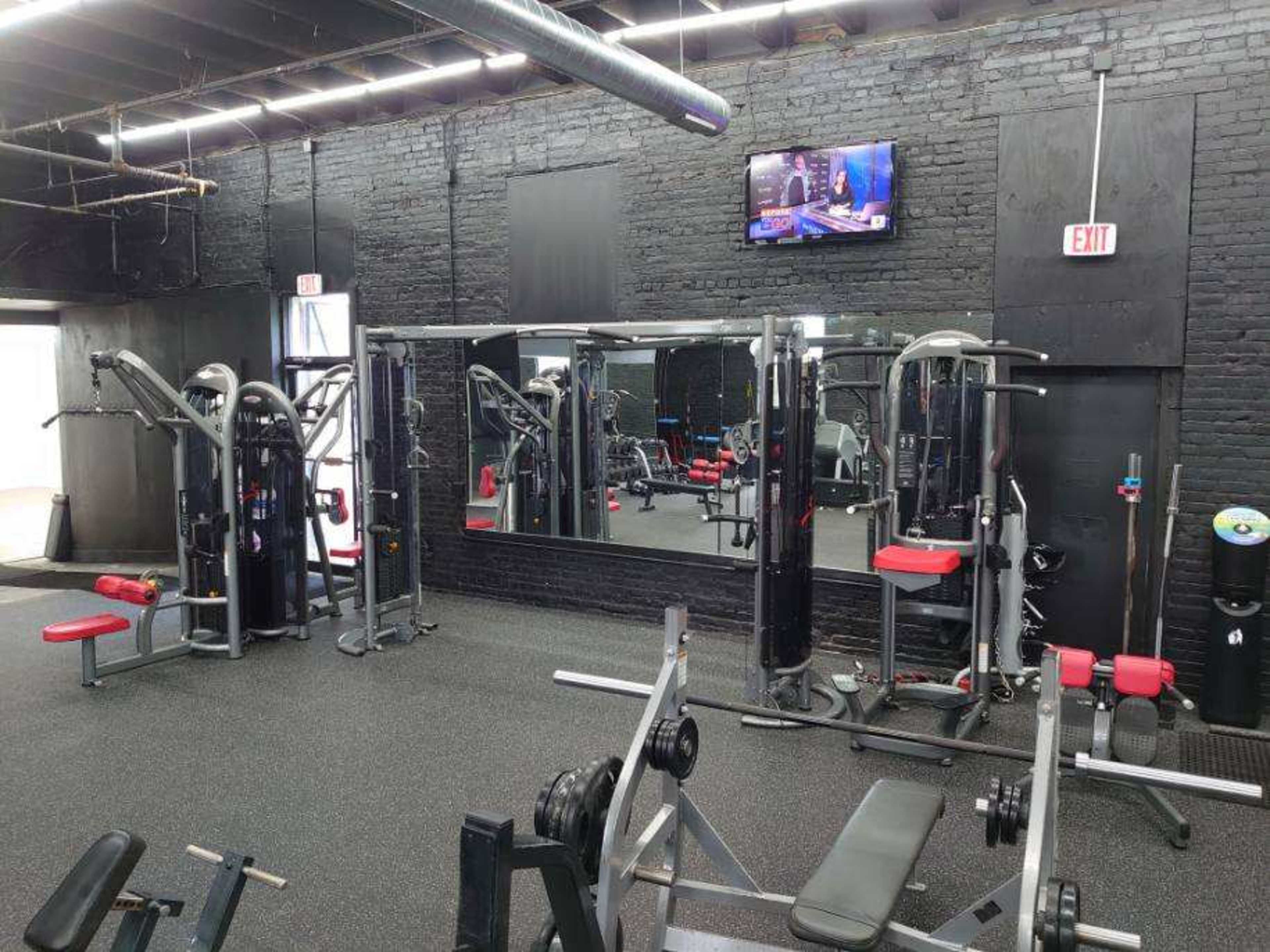The image shows a gym interior with workout equipment, including weight machines and benches, set against a black brick wall, and a television mounted above.