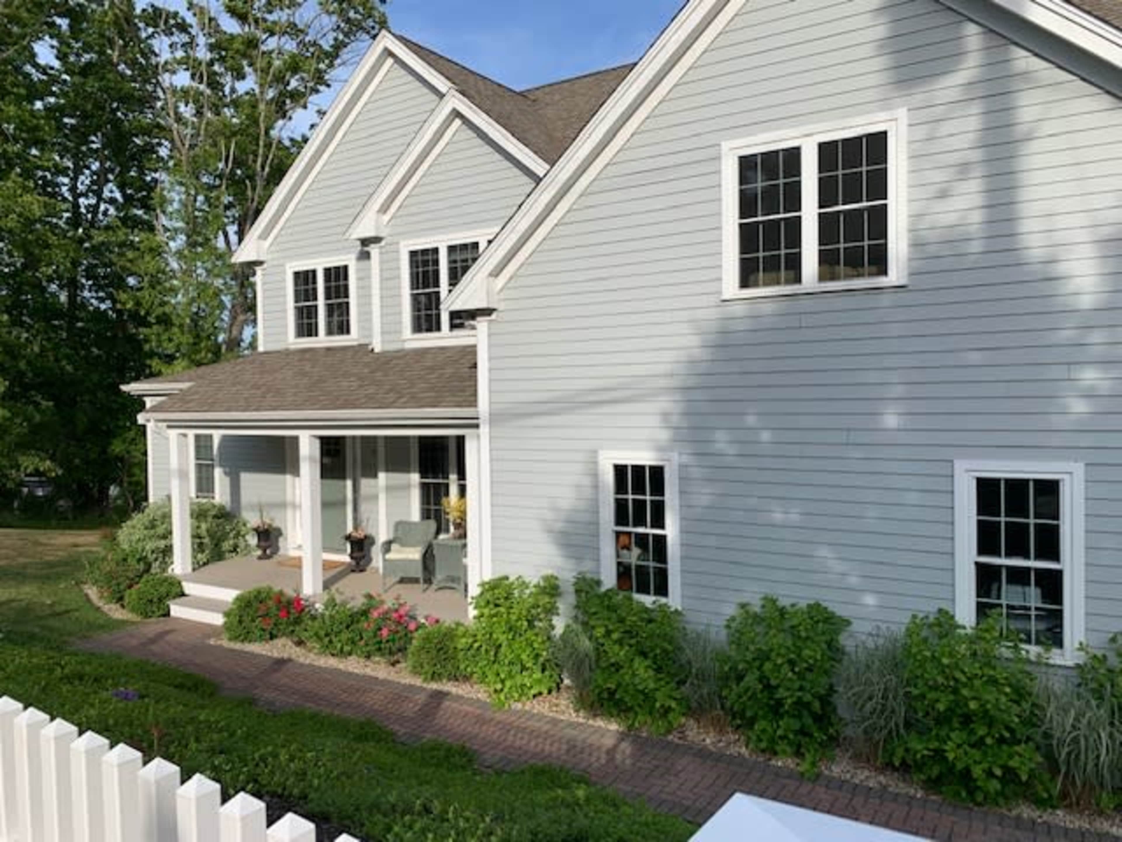 A light blue house with multiple windows and a porch is surrounded by greenery and blooming flowers.