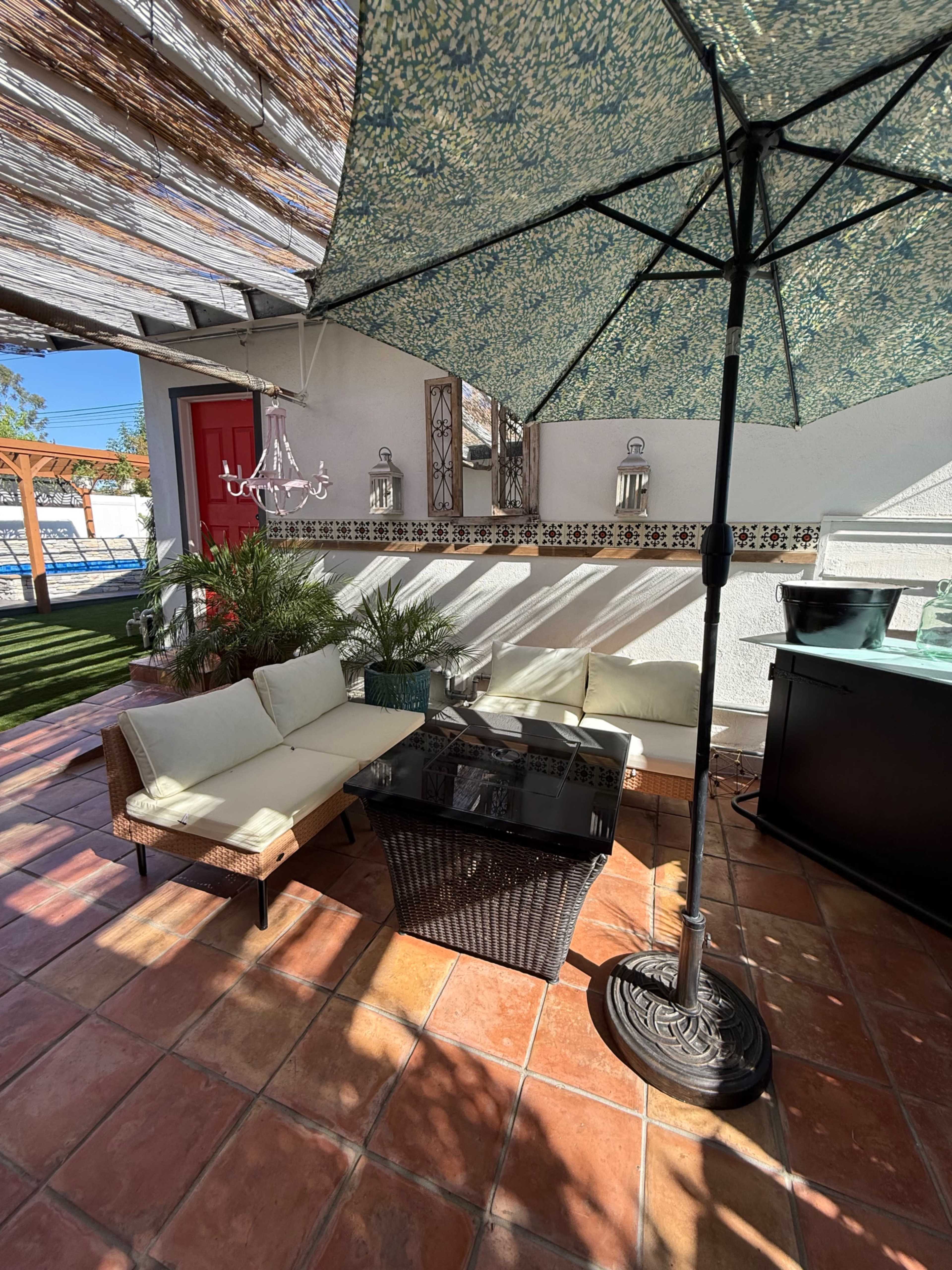 A shaded outdoor seating area features a wicker table and sofa set under a large umbrella on a tiled patio.
